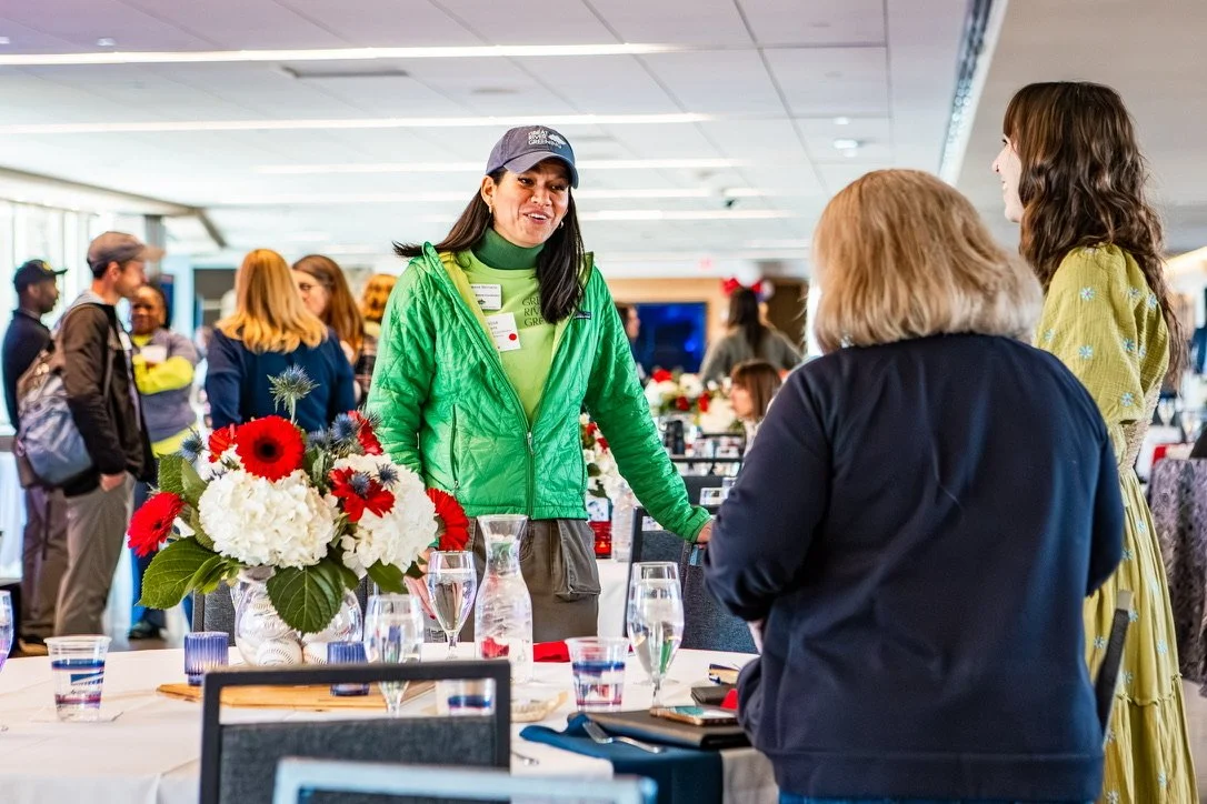 A woman in a green jacket and gray cap speaking to two women at a table with a floral centerpiece, with several people in the background at an indoor event.