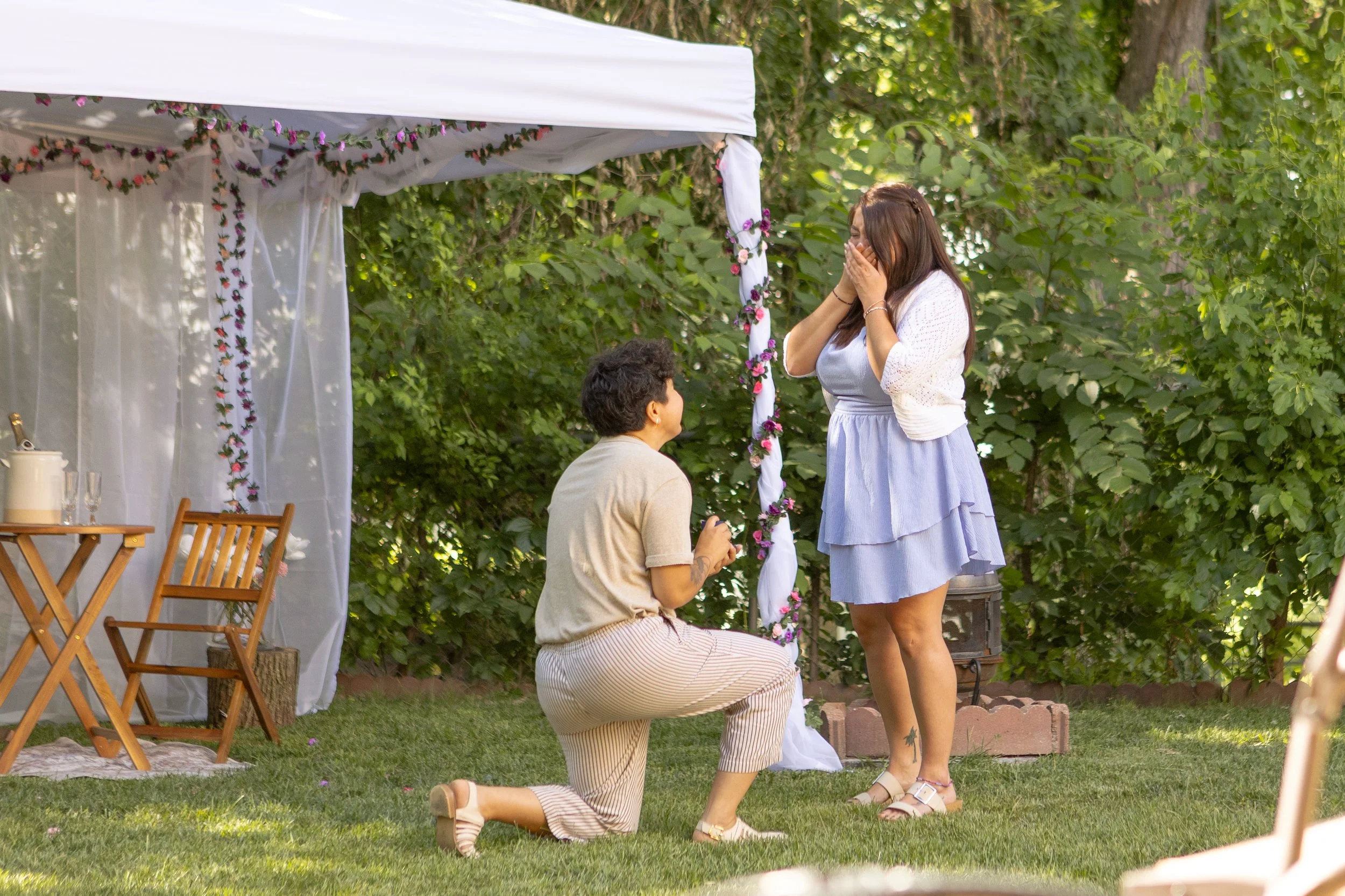 A woman proposing to a woman kneeling on one knee outdoors, with a woman covering her mouth in surprise. There is a decorated wedding arch in the background and surrounding trees.