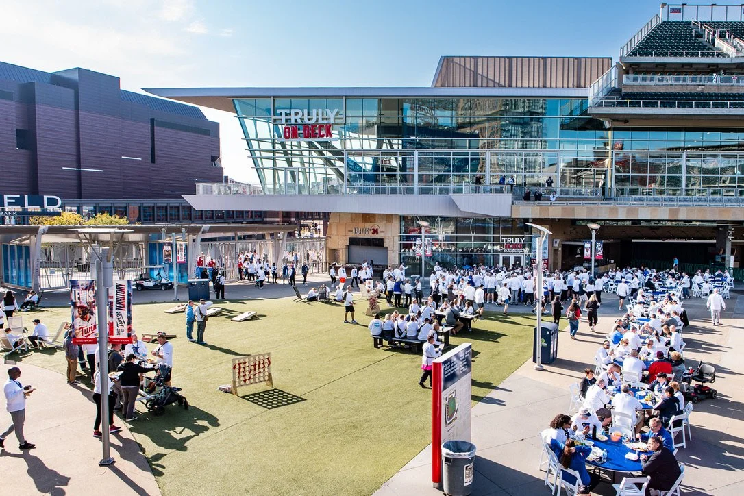 Outdoor patio area outside a sports stadium with numerous people dining and socializing, some in white shirts, under clear skies.