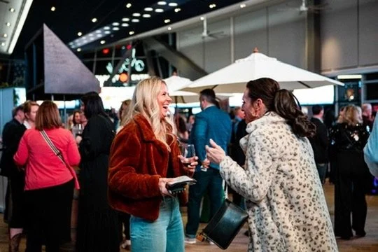 Two women are talking and laughing at a social event or party, with other people mingling in the background.