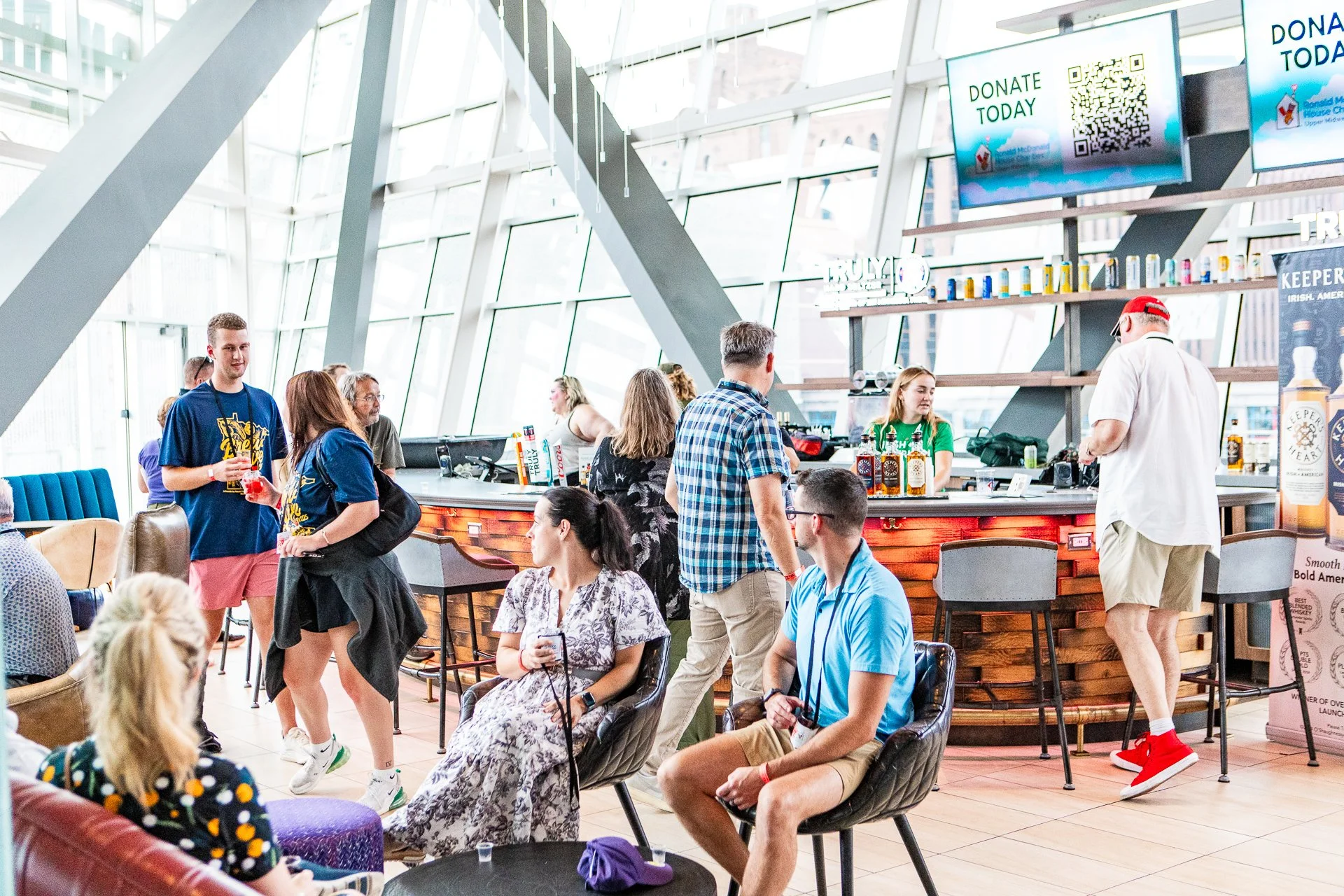 People gathered in a modern, spacious indoor area with a bar and large screens displaying 'Donate Today' and a QR code, suggesting a charity or fundraising event.