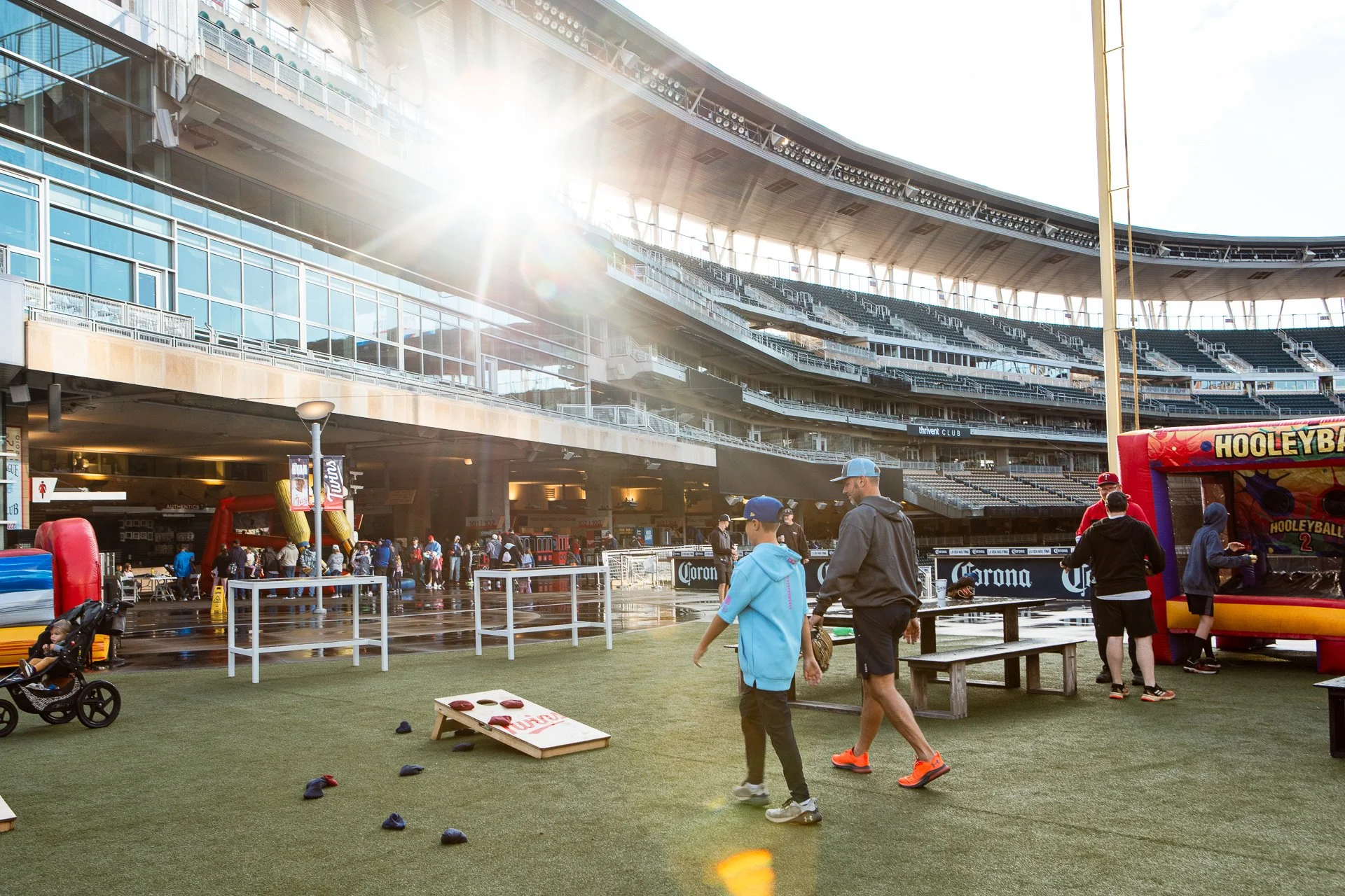 People enjoying activities in a stadium concourse area with a large seating section overhead, sunlight shining through, and a Hooley Volleyball attraction.