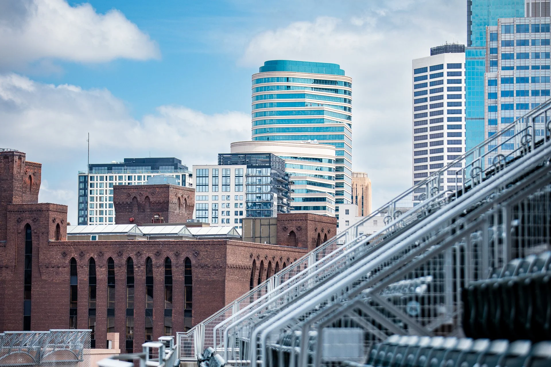 The Minneapolis skyline with modern glass skyscrapers and older brick buildings, viewed from a stadium with empty seats and metal railings in the foreground.