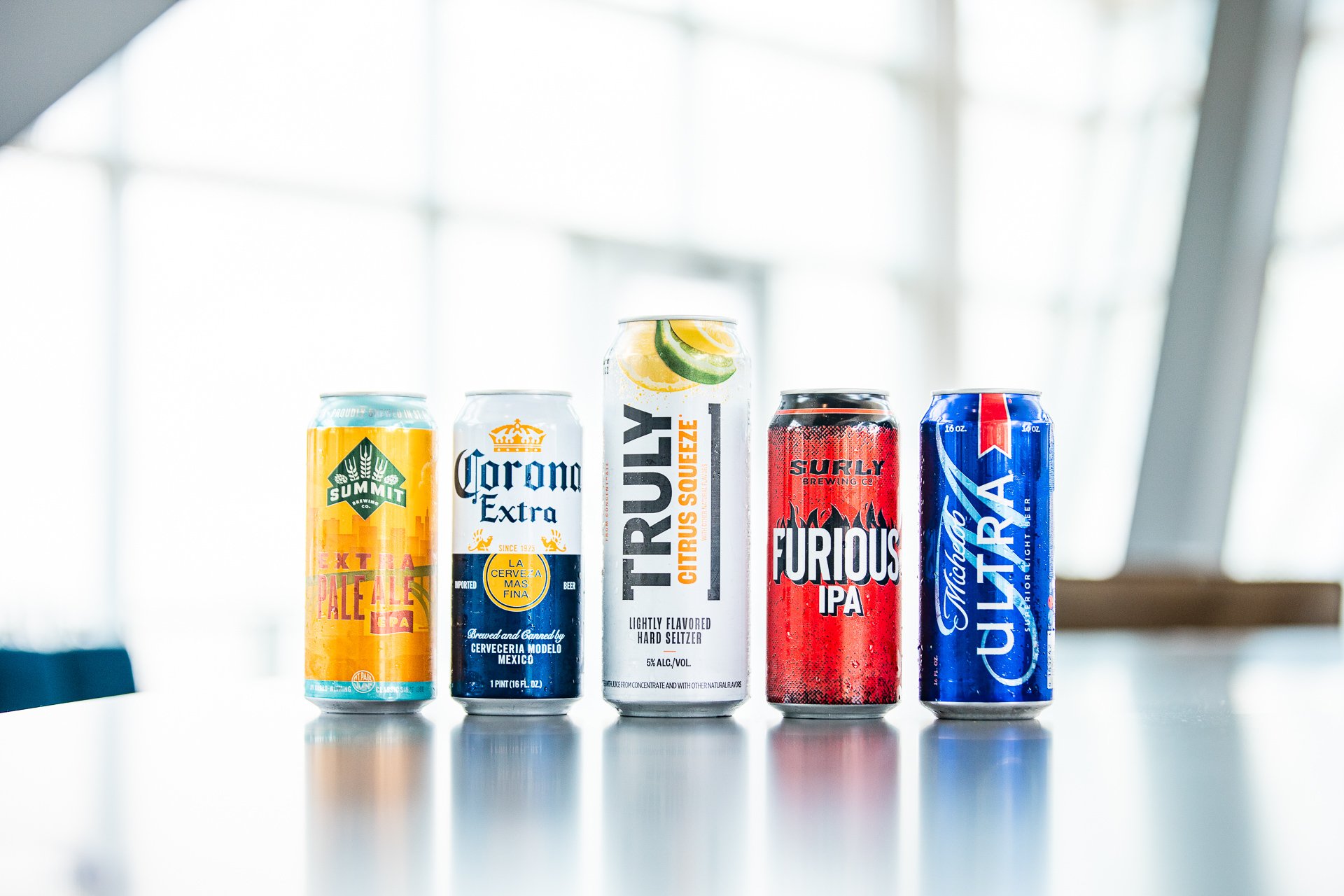 Five cans of beer and hard seltzer beverages displayed on a table with a bright background. The cans feature colorful and bold labels with names like Summit Extra Pale Ale, Corona Extra, Trusy Citrus Squeeze, Furious IPA, and Uinta Lucky Lite.