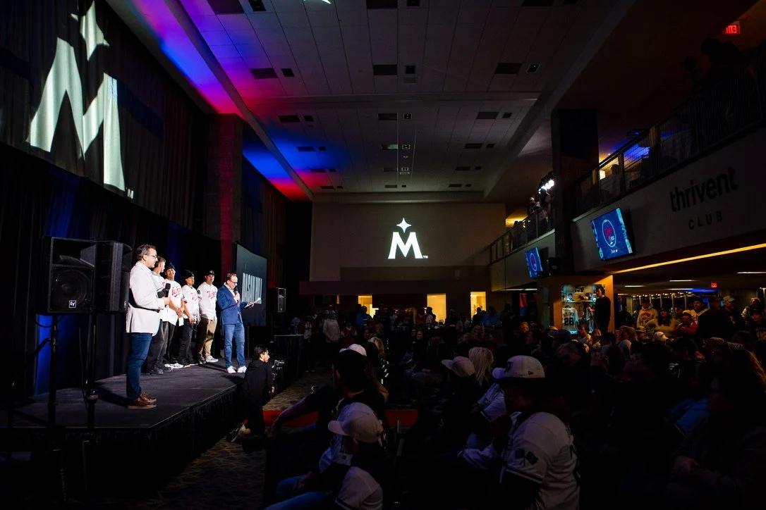 A group of people on stage at an event, with a large screen behind displaying a star and the letter 'M'. Audience members seated and standing in a dark,ly lit room.