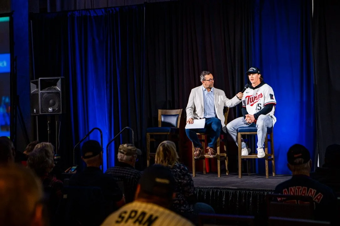A woman in a baseball jersey being interviewed on stage by a man in a light gray blazer, with an audience watching.