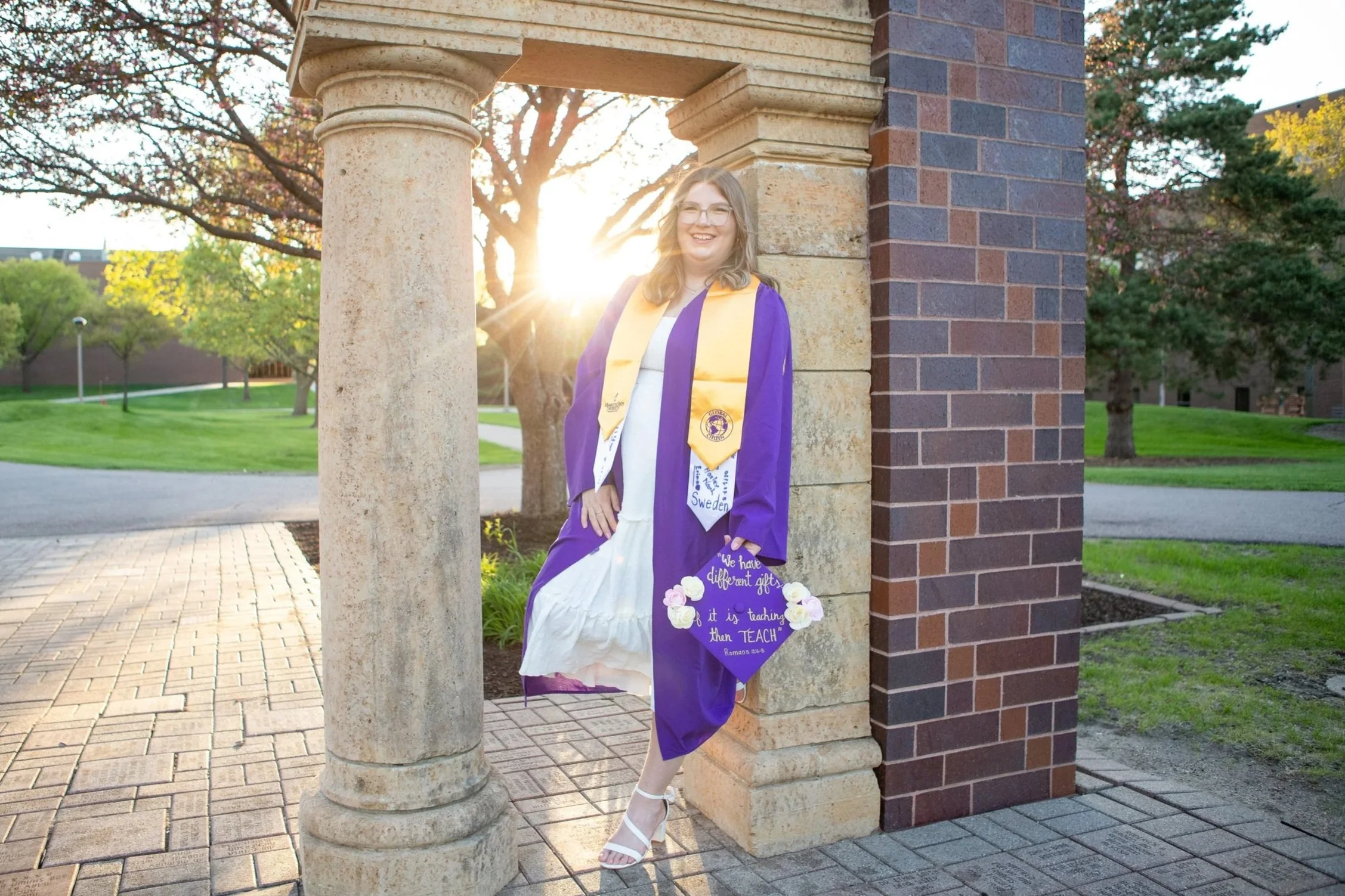 A young woman in a graduation gown and cap, smiling, standing under a stone archway with the setting sun behind her, in a park with trees and grass.