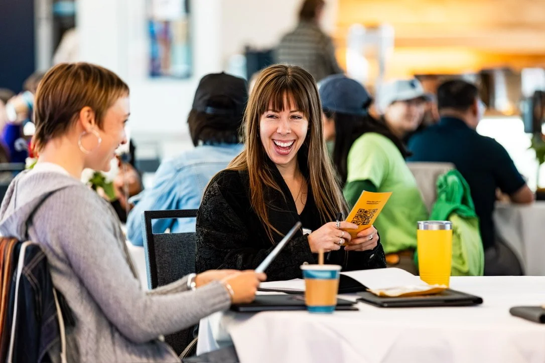 Two women sitting at a table in a busy café, laughing and chatting, with drinks and notebooks in front of them.