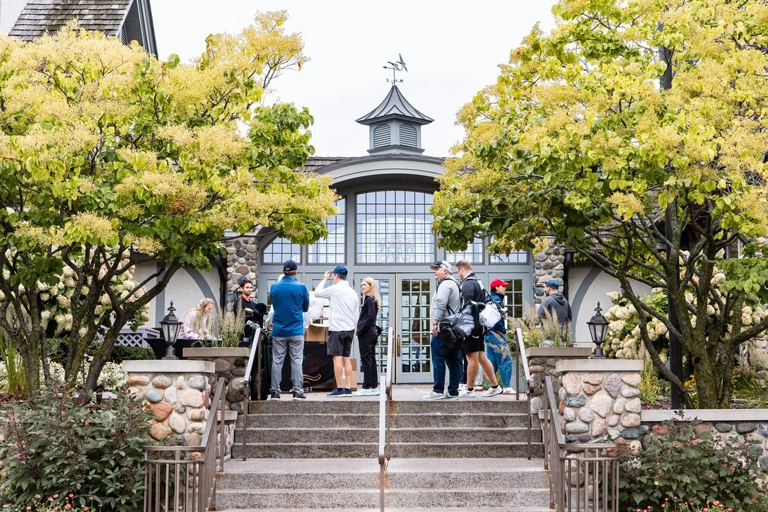 Group of people gathering outside a building with large glass doors, surrounded by trees and stone walls, during daytime.