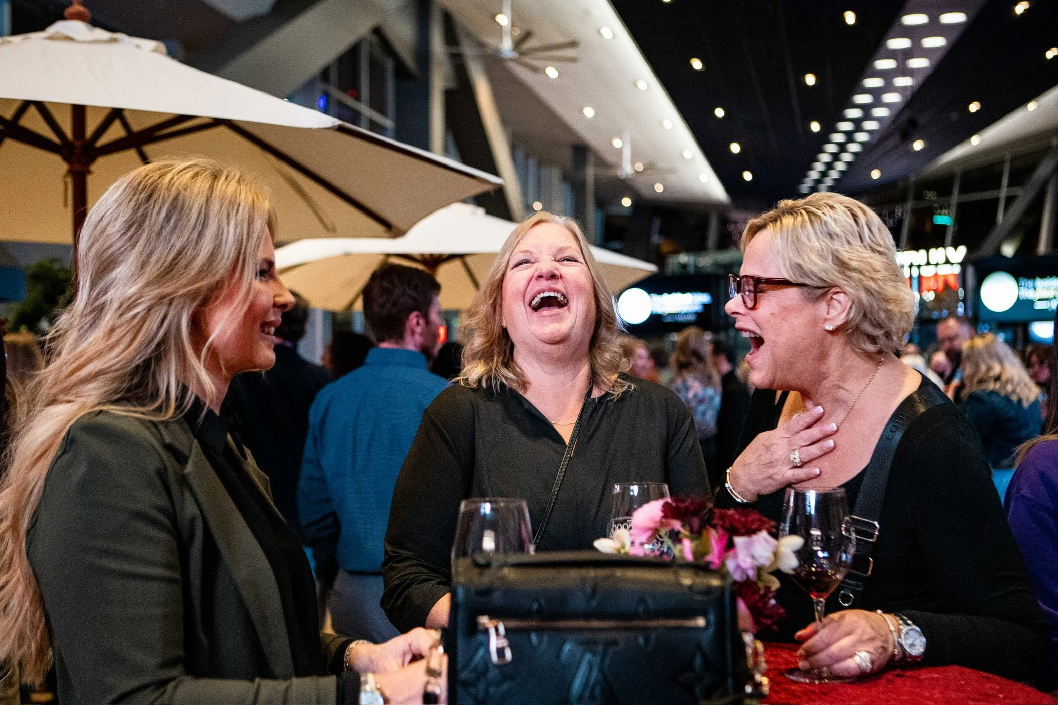 Three women laughing and having a conversation at a social event or restaurant, with glasses of wine and flowers on the table, in a modern indoor setting.