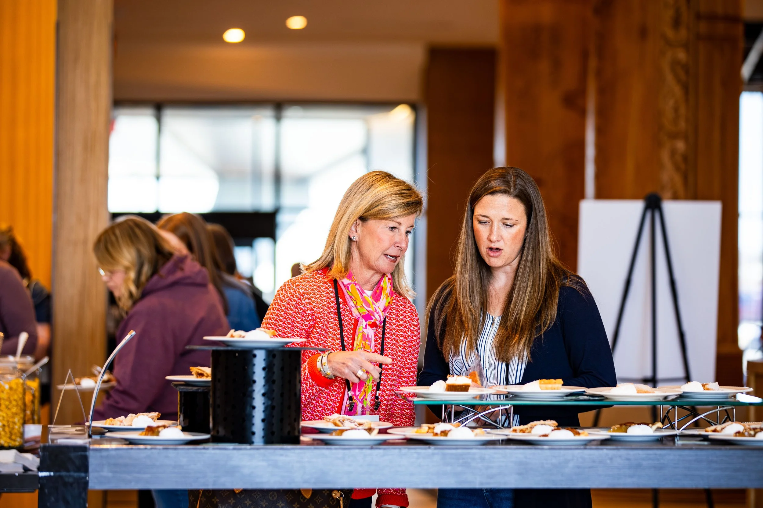 Two women looking at food on a buffet table at a social event or conference.