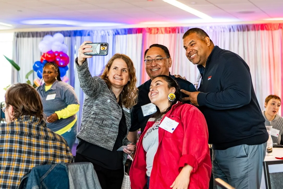 A group of four people smiling and taking a selfie at an indoor event with colorful balloons and drapes in the background.
