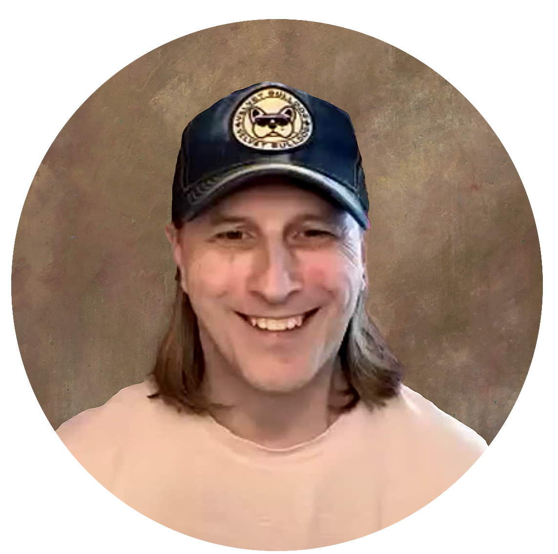 Smiling man with long hair wearing a black baseball cap with a patch of a dog wearing sunglasses, beige shirt, and a brown textured background.
