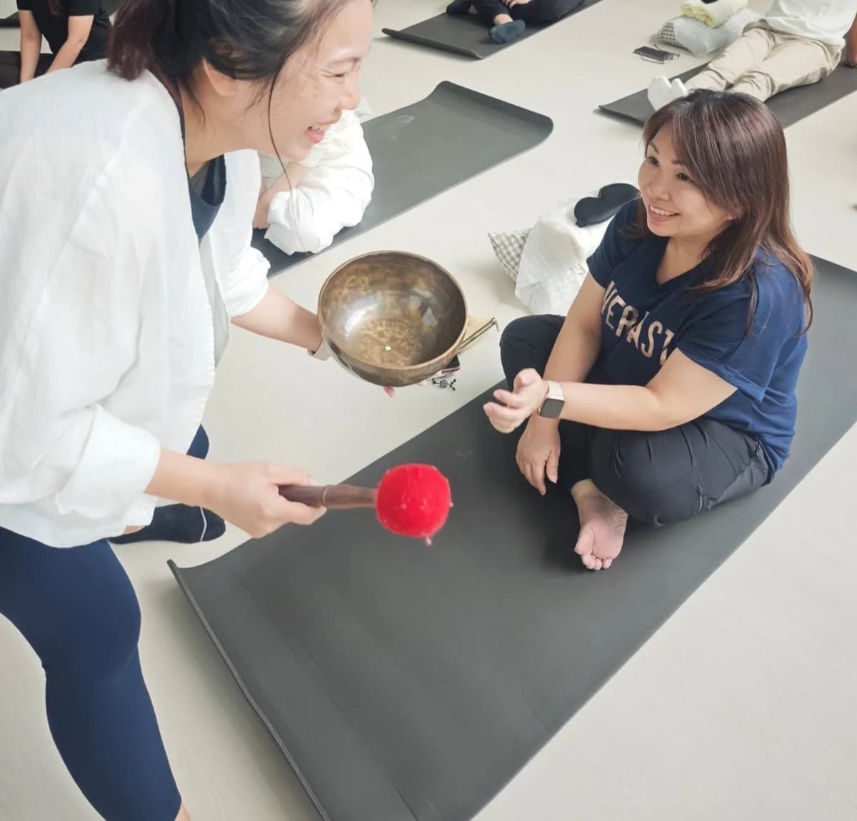 Group of people participating in a sound healing or meditation session indoors, with singing bowls, candles, and decorative items on a wooden floor.