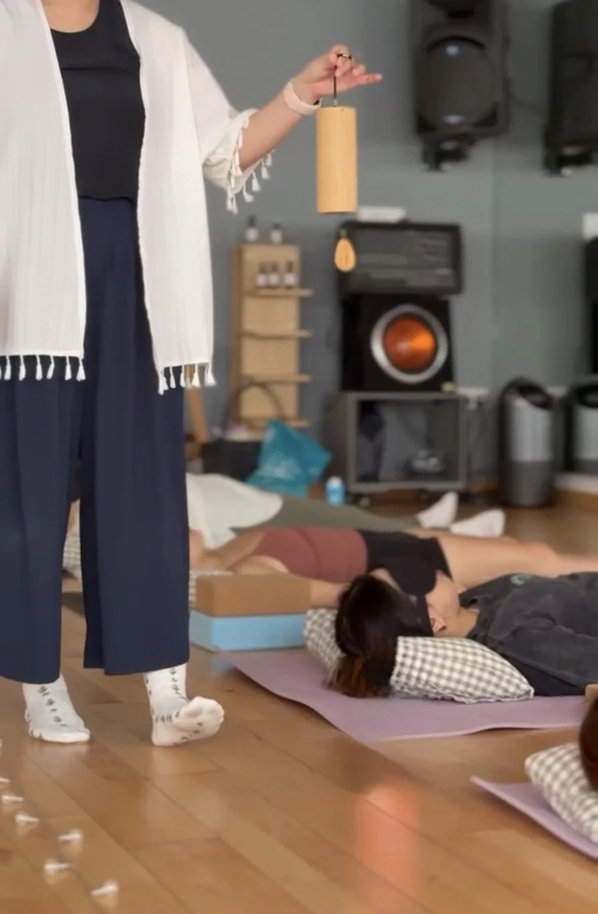 People lying on mats with eyes covered, participating in a guided meditation session led by a person standing near a window with light filtering through closed blinds.