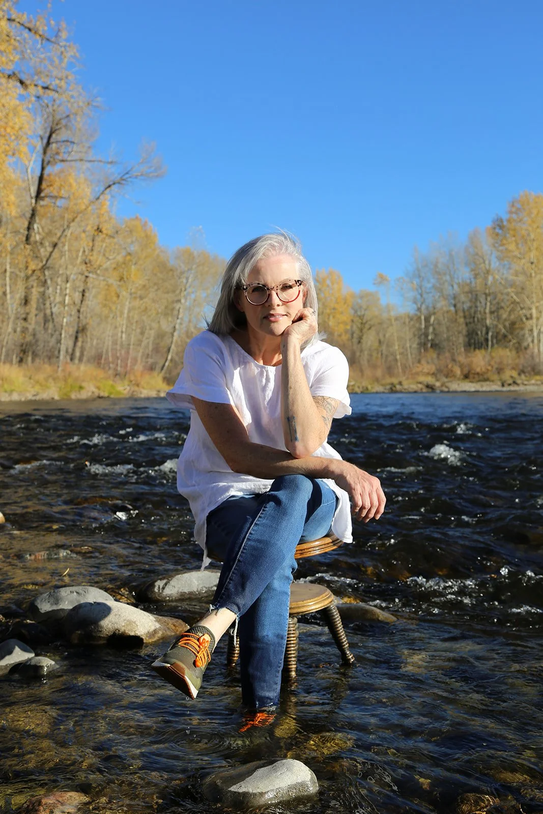 A woman with gray hair and glasses sitting on a small chair in a shallow river, with trees and blue sky in the background.