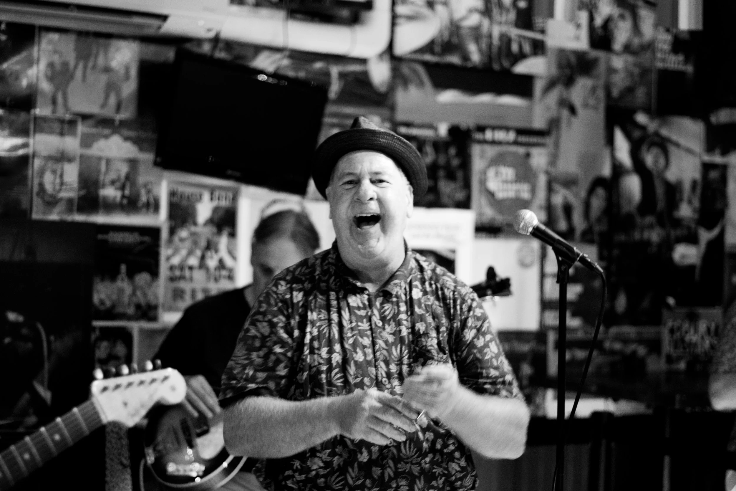 A man singing with his eyes closed, wearing a hat and floral shirt, in a music venue with posters and a microphone in front of him.