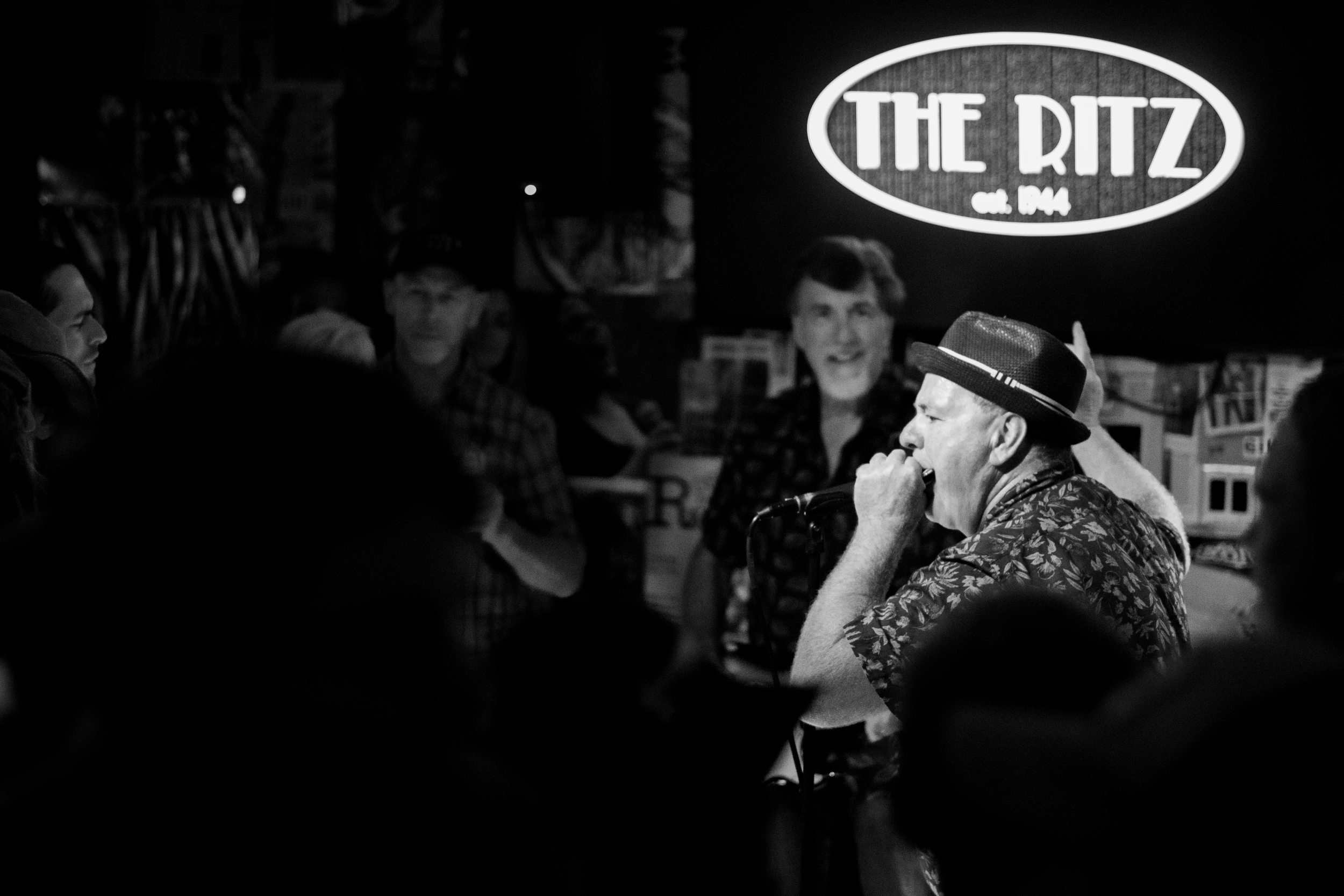 A man wearing a fedora hat and floral shirt singing into a microphone on a stage, with the sign 'The Ritz est. 2014' illuminated in the background, and an audience watching.