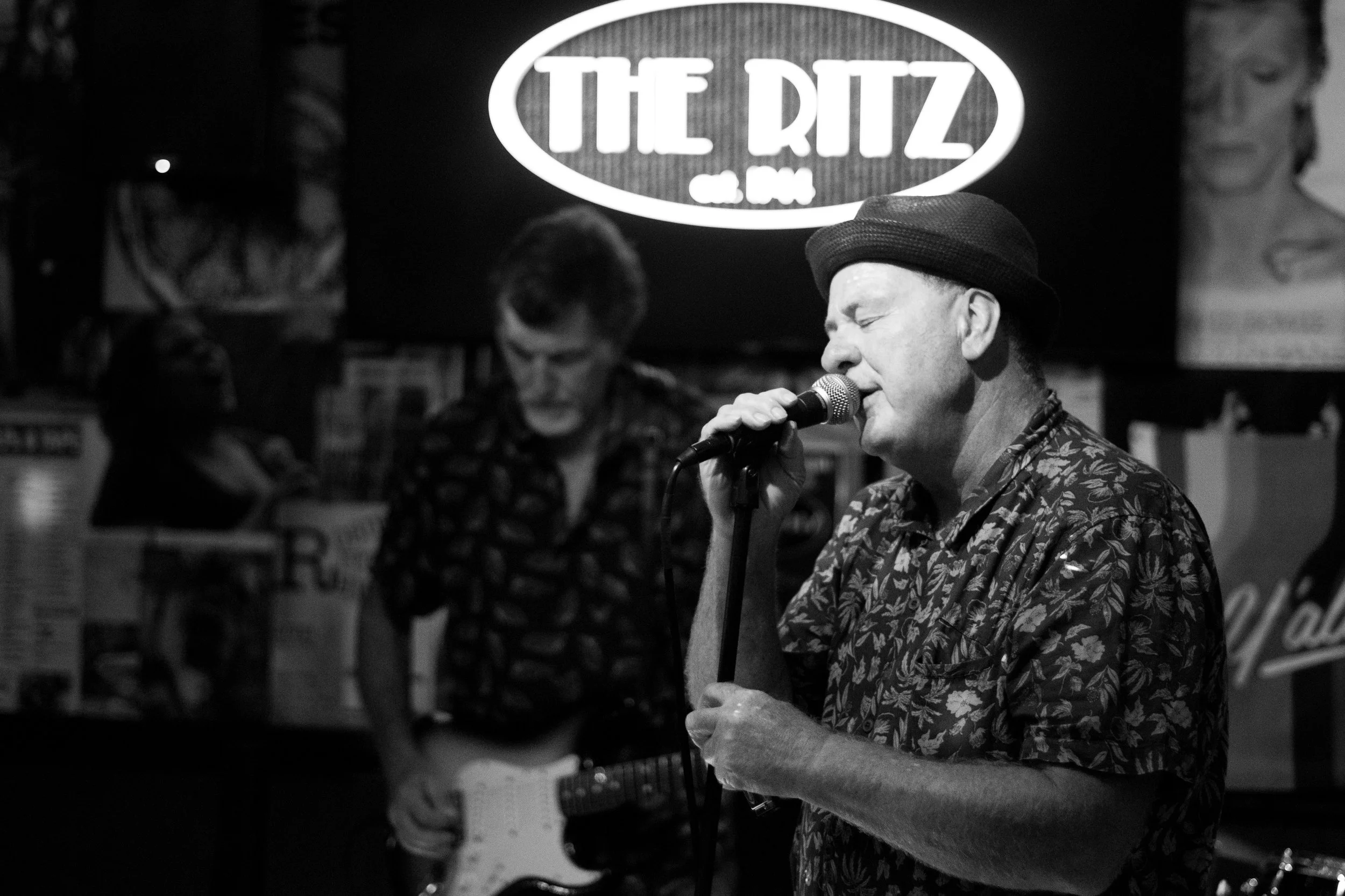 A man singing into a microphone with eyes closed at a live music venue, with a neon sign that says 'The Ritz' in the background and another man playing guitar behind him.