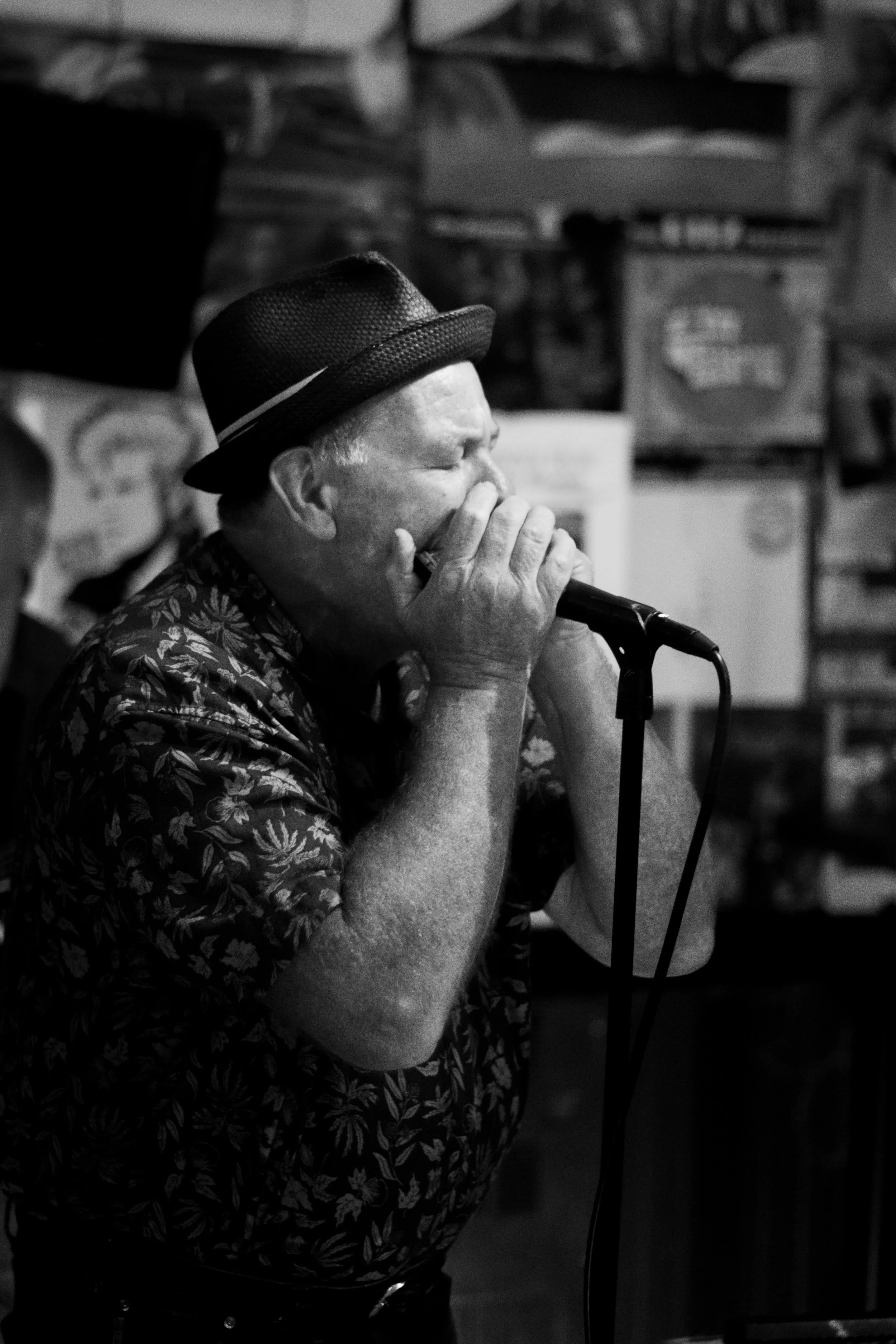 A man wearing a hat and floral shirt playing a harmonica into a microphone in a dimly lit setting.