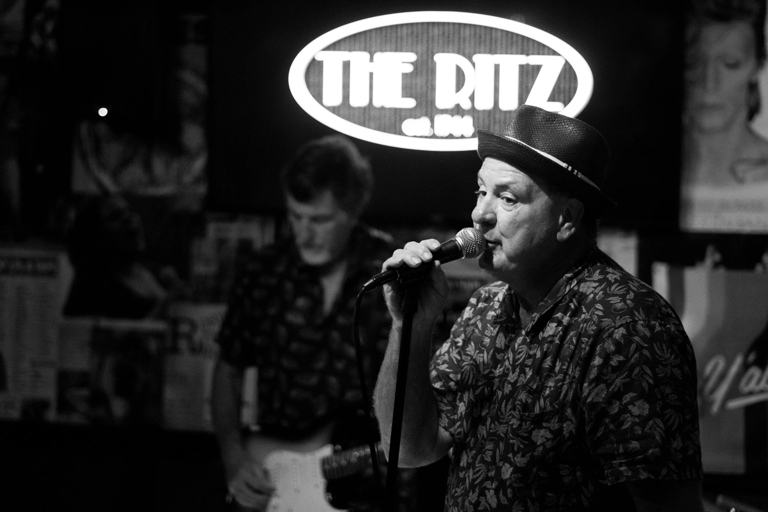 Black and white photo of a man singing into a microphone at a bar or music venue, with a person playing guitar in the background, and a lit sign that reads "The Ritz" behind them.