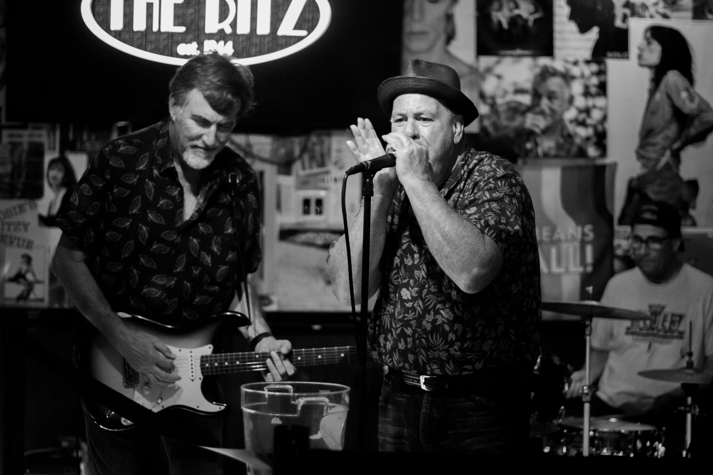 Black and white photo of a band performing in a bar or club. A man with a fedora hat sings into a microphone while another man plays an electric guitar. A drummer is visible in the background. Posters and photographs are on the wall behind them.