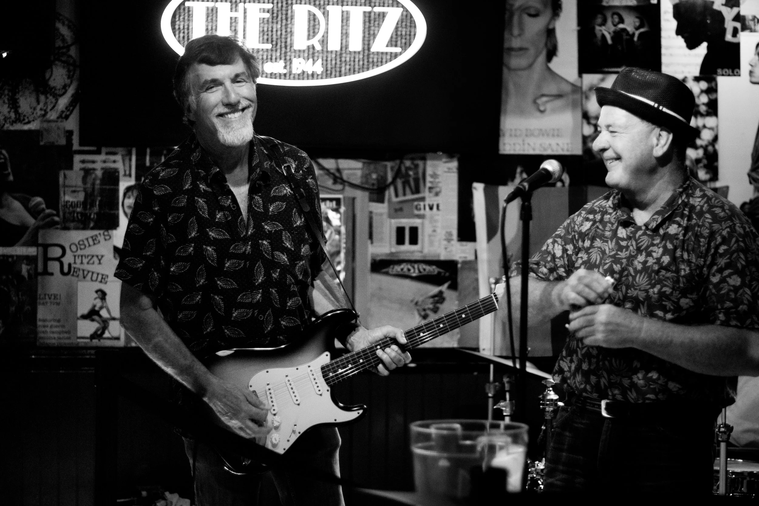 Two men smiling and enjoying music performance at a live music venue, one holding an electric guitar, with posters and a neon sign for 'The Ritz' in the background, in black and white.