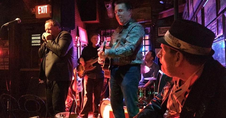 A live band performs in a dimly lit bar or club, with a lead singer, guitarist, bassist, and drummer. The scene has warm, colorful lighting and a cozy, intimate atmosphere.