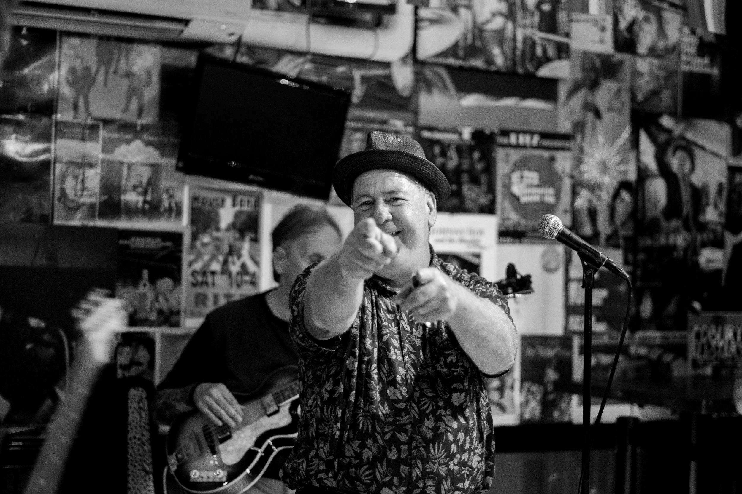 A man in a floral shirt and fedora hat pointing at the camera with a smile, standing in front of a microphone, with a person playing guitar behind him.
