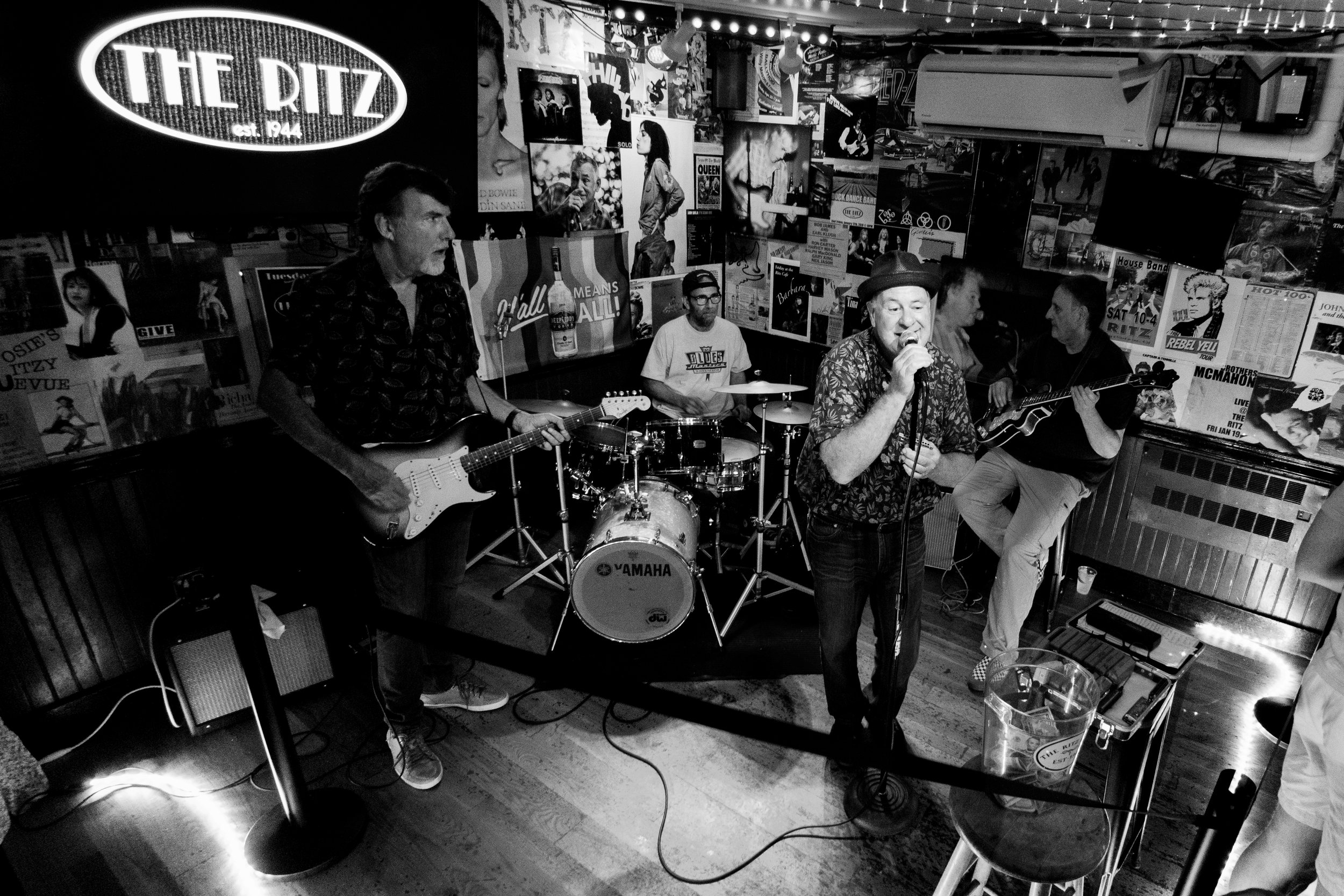A band performing live at a bar named The Ritz, with four musicians playing electric guitar, drums, and singing. The bar walls are decorated with posters and artwork.