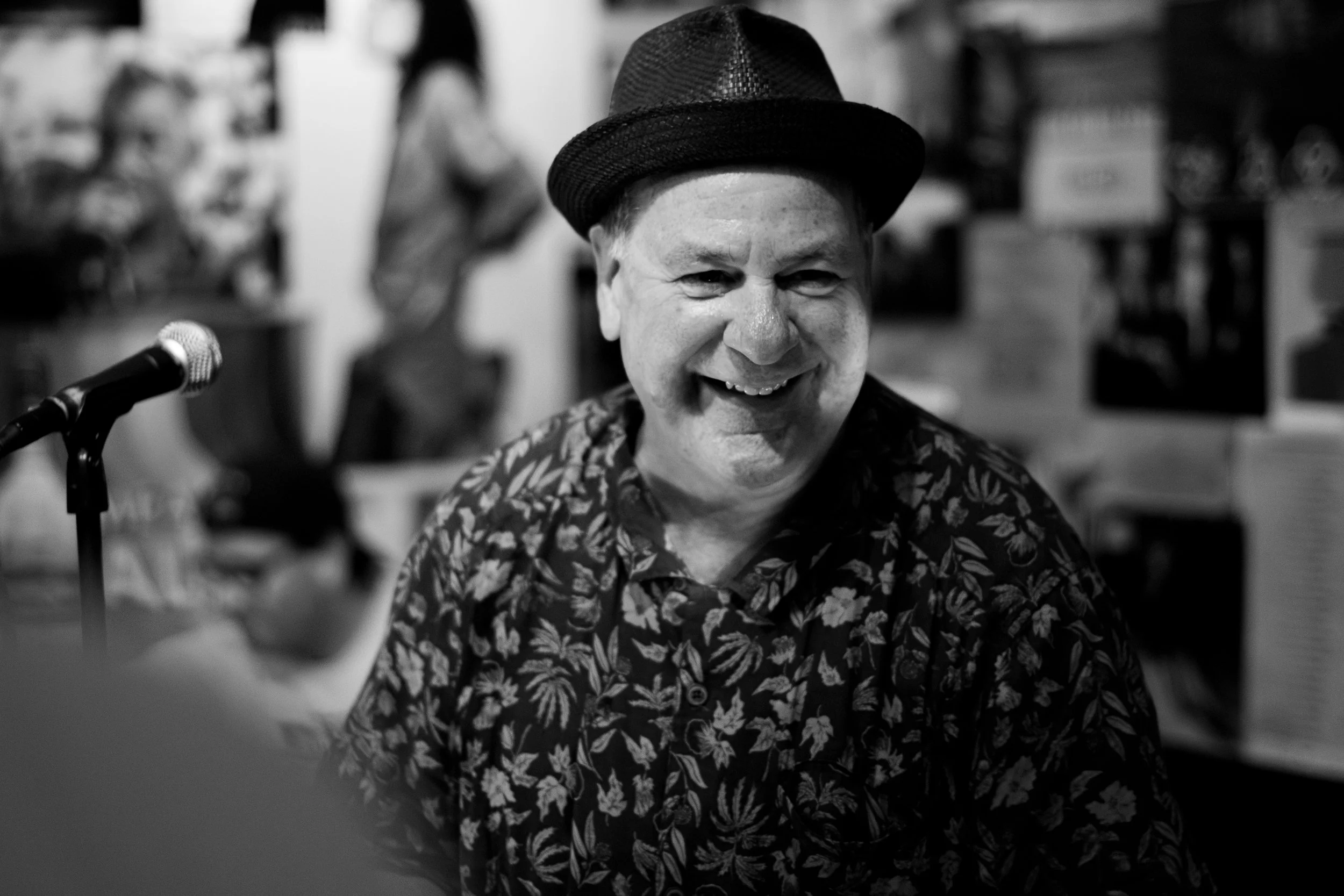 A smiling man wearing a hat and patterned shirt, sitting in a room with blurred artwork and a microphone nearby.