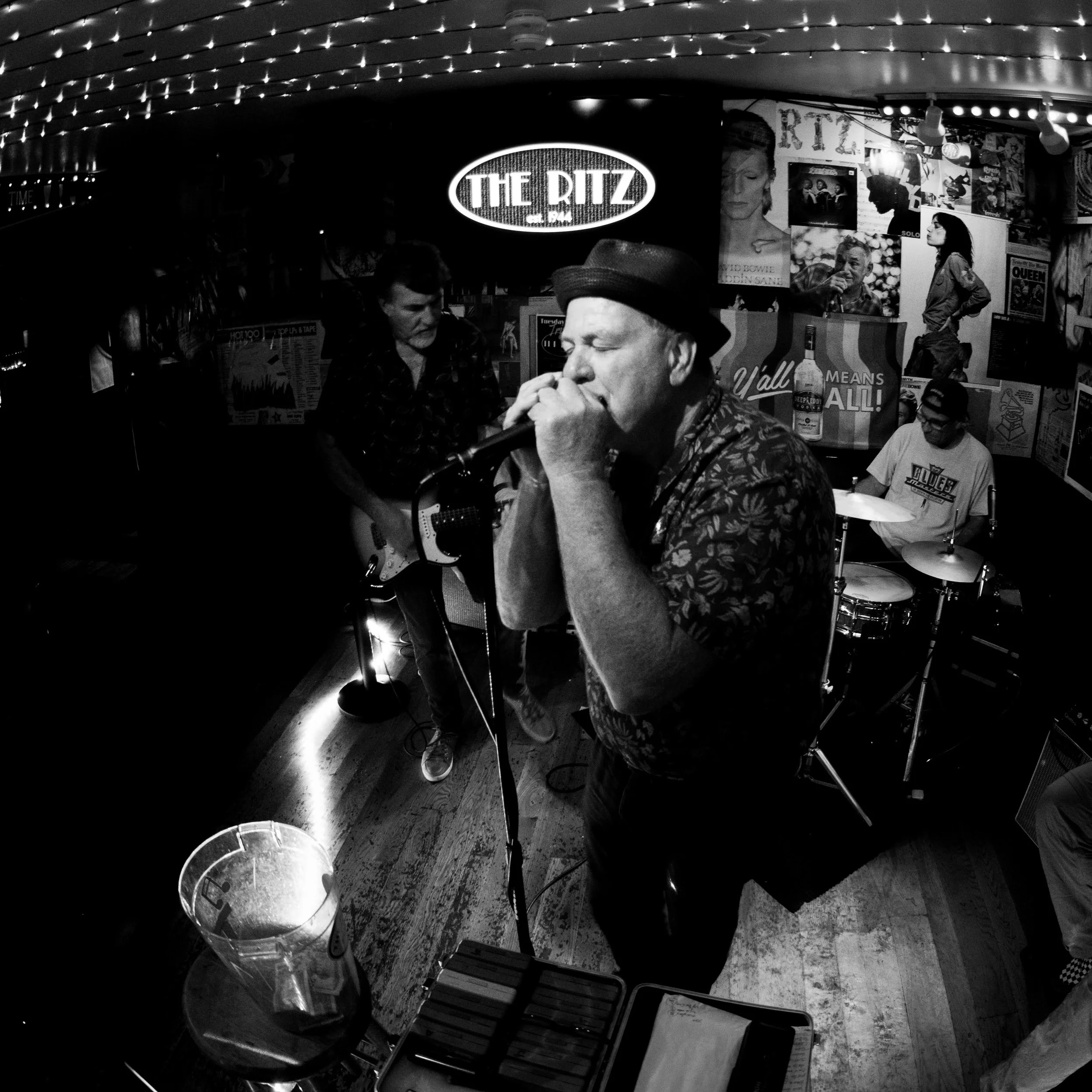 A black and white photo of a band performing live in a small, decorated venue. The lead singer is singing into a microphone, wearing a hat and a floral shirt. Behind him, a guitarist and a drummer are visible, with posters and signs on the wall, incl