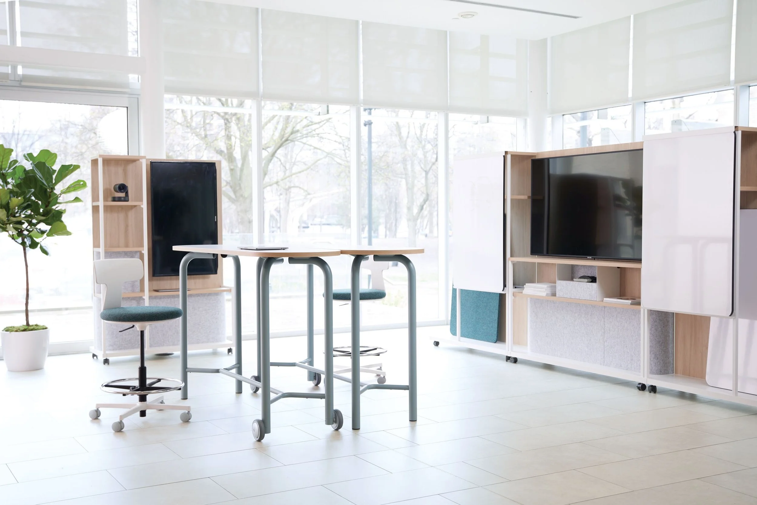 Modern office space with white walls, large windows, a small round table with two chairs, a potted plant, and a large flat-screen TV on a wooden stand.