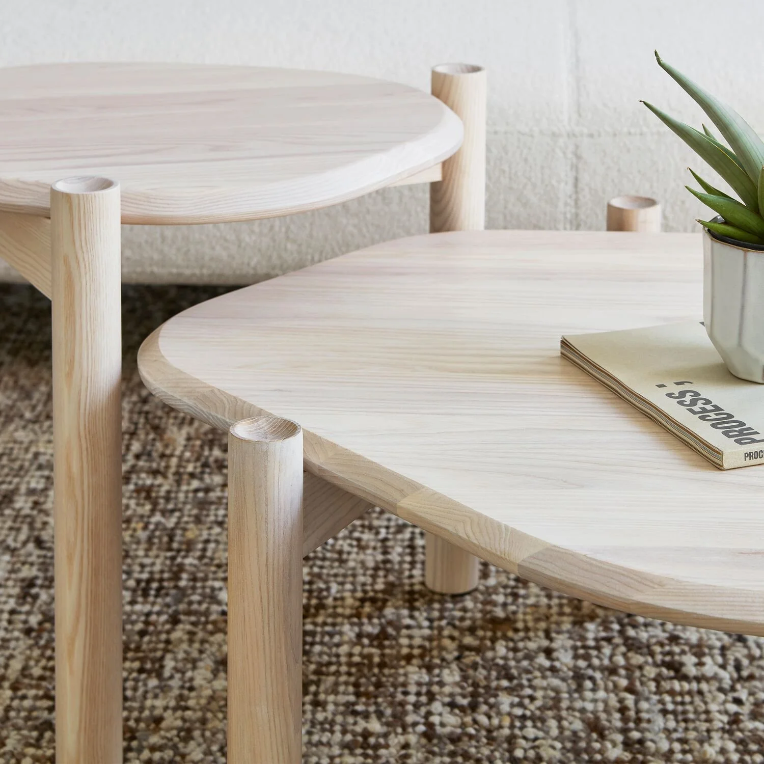 Two minimalist wooden tables with rounded edges, a potted plant, and a magazine on a textured rug near a white wall.
