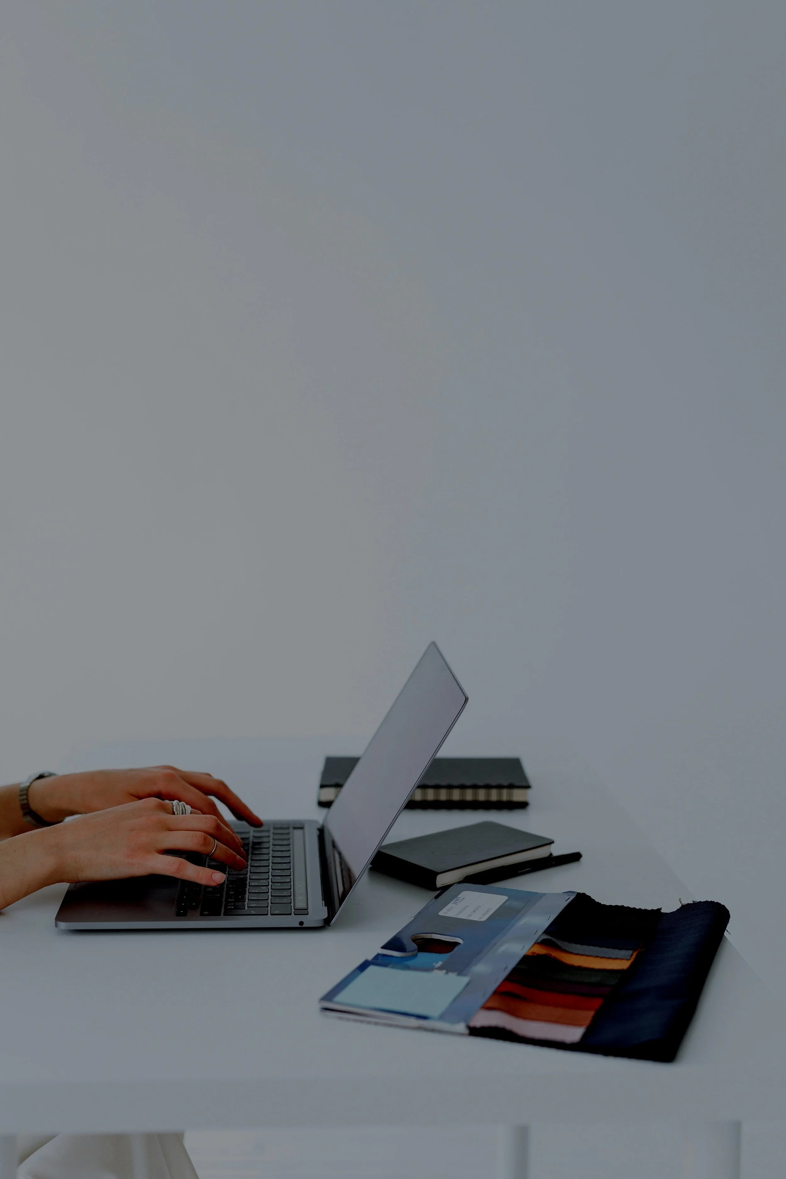 Person typing on a laptop at a desk with notebooks and fabric samples nearby at Authent.