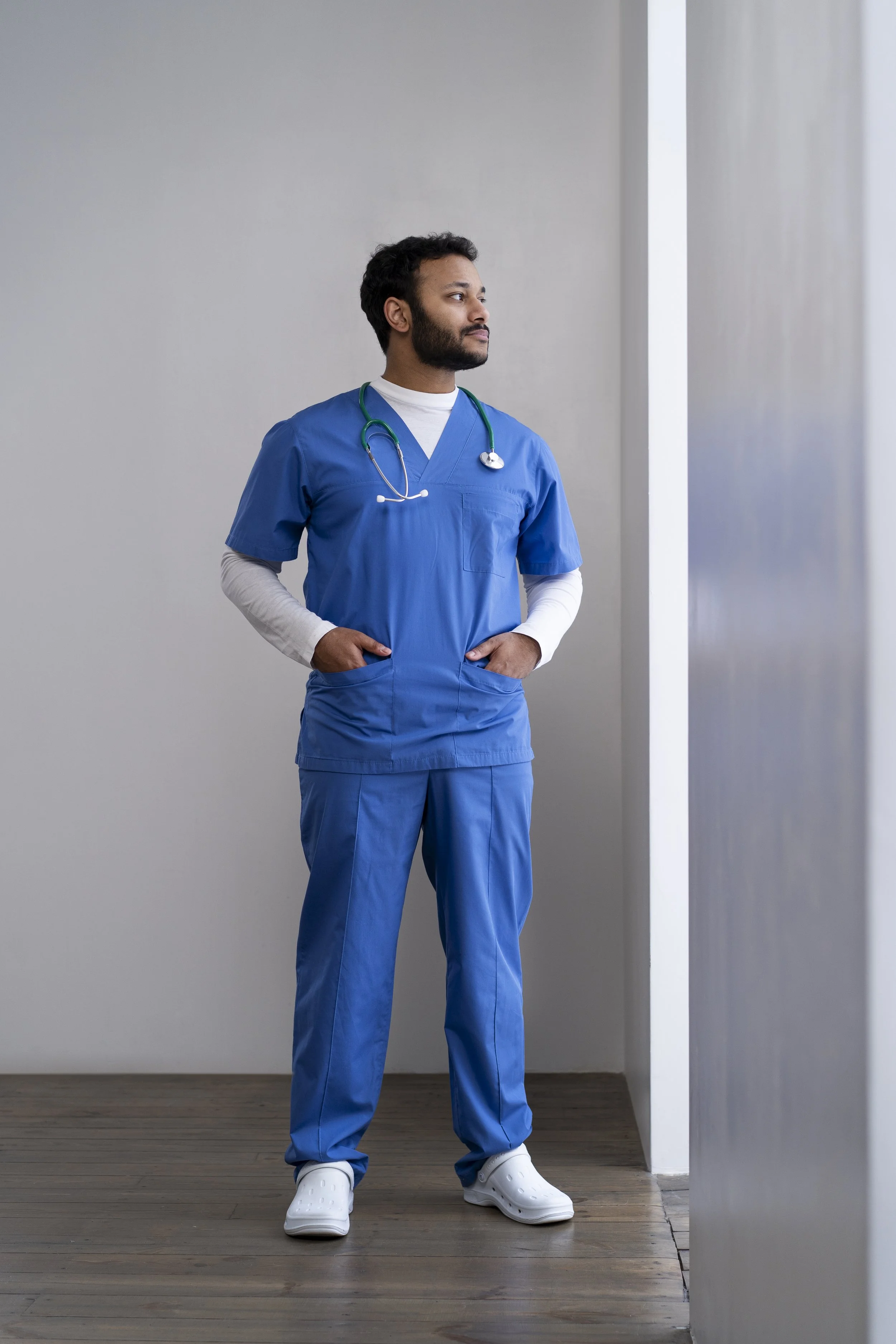 A healthcare professional in blue scrubs manufactured at Authent standing with hands in pockets, looking out a window.