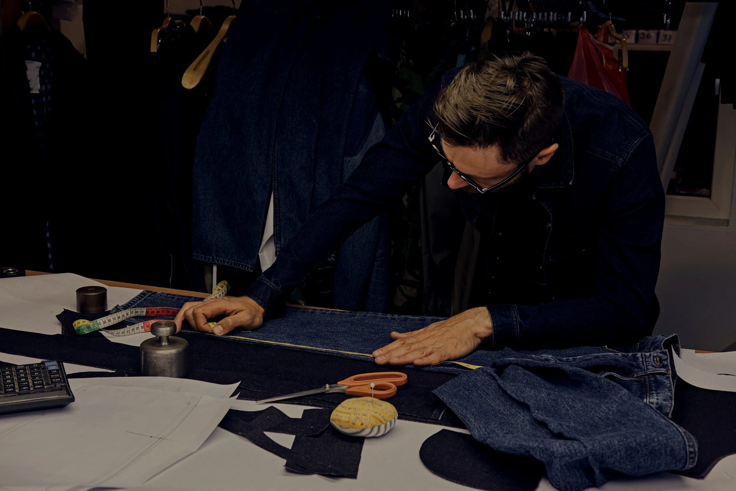 Tailor measuring denim fabric with measuring tape, surrounded by scissors, pattern papers, and sewing tools for accuracy at Authent office.