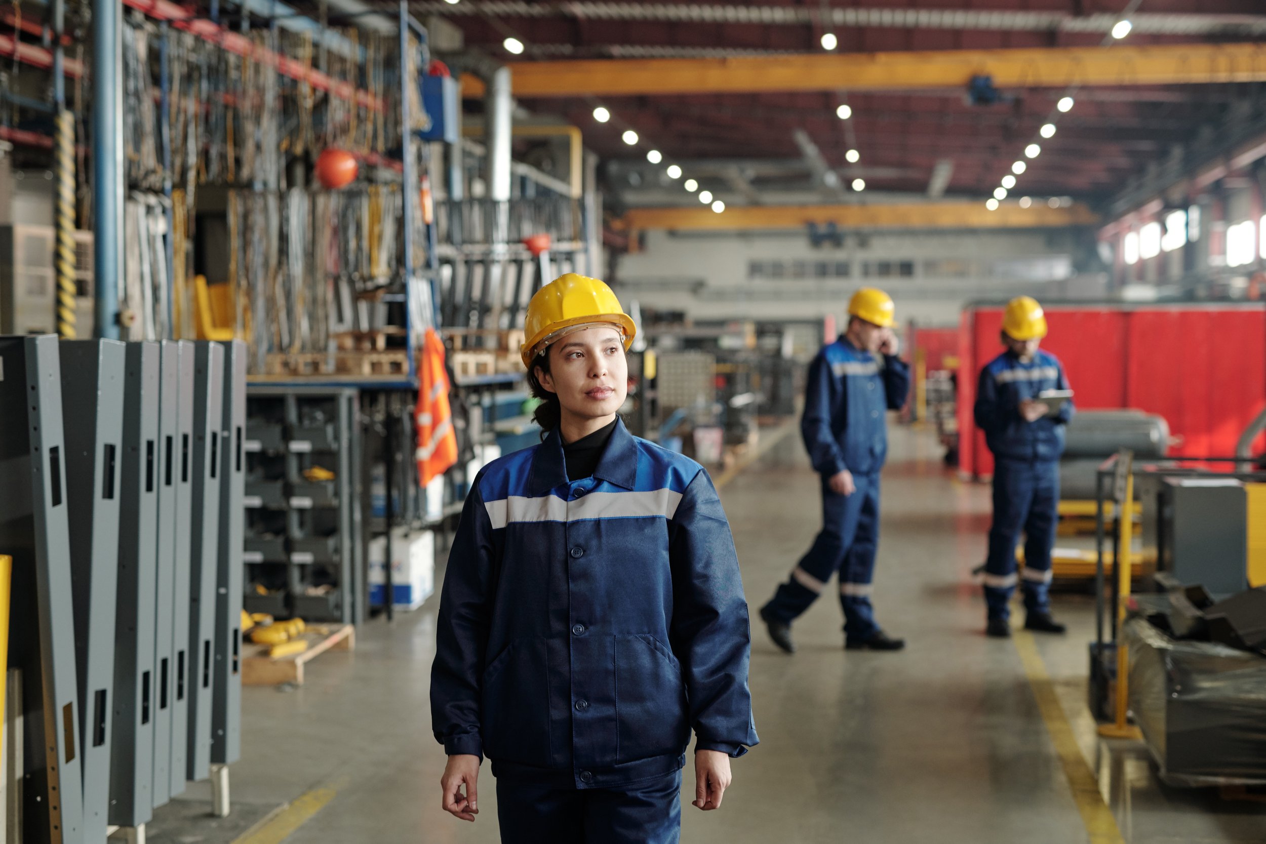 A woman in a blue reflective and protective uniform and yellow hard hat stands in a factory setting, with two other workers in similar attire - All maufactured at Authent. 