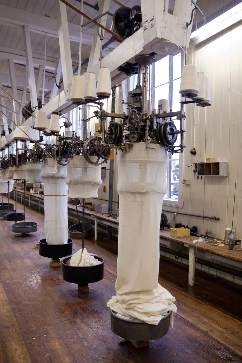 Vintage sock knitting machines in a textile factory setting, with large cone yarn spools above and knitted fabric hanging below.