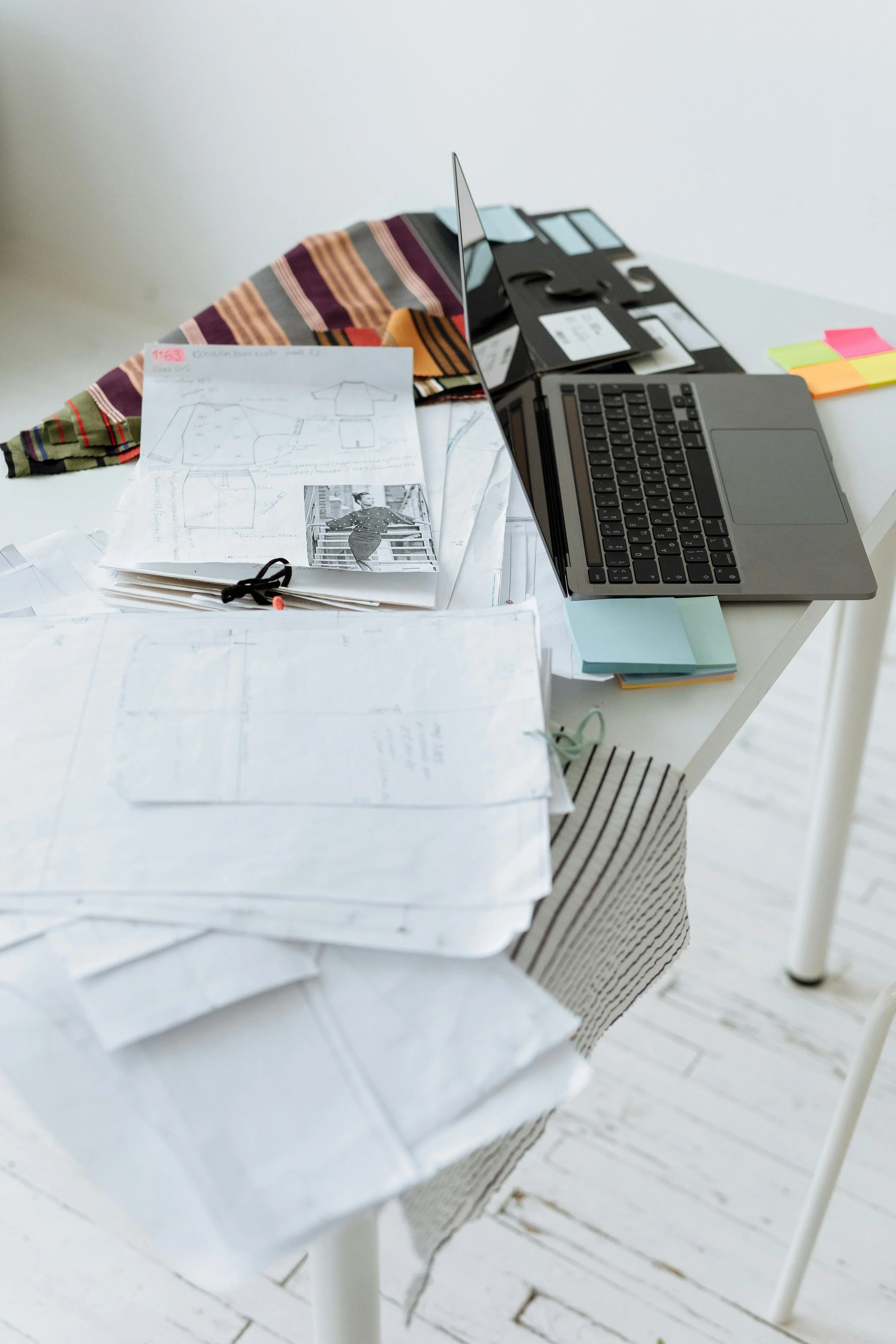 Cluttered workspace with laptop, sewing patterns, fabric swatches, and sticky notes on a white desk at Authent Office.