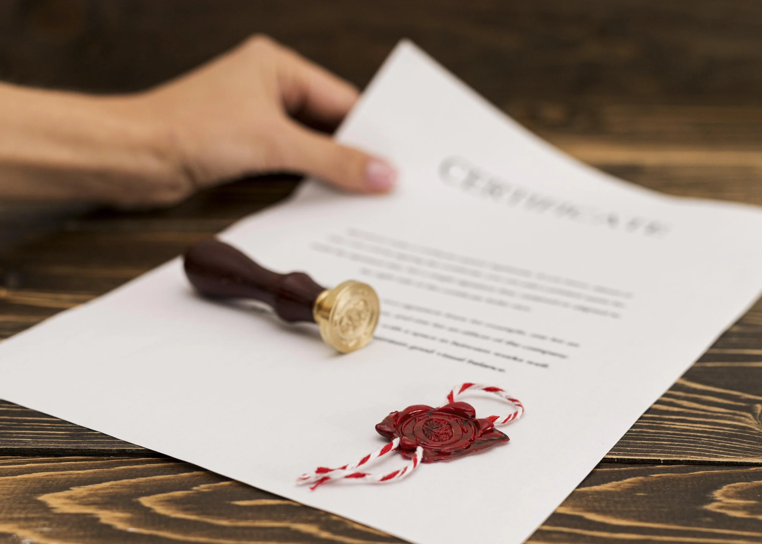 A hand holding a certificate with a red wax seal and a stamp resting on the table. Certification and clients of Authent.