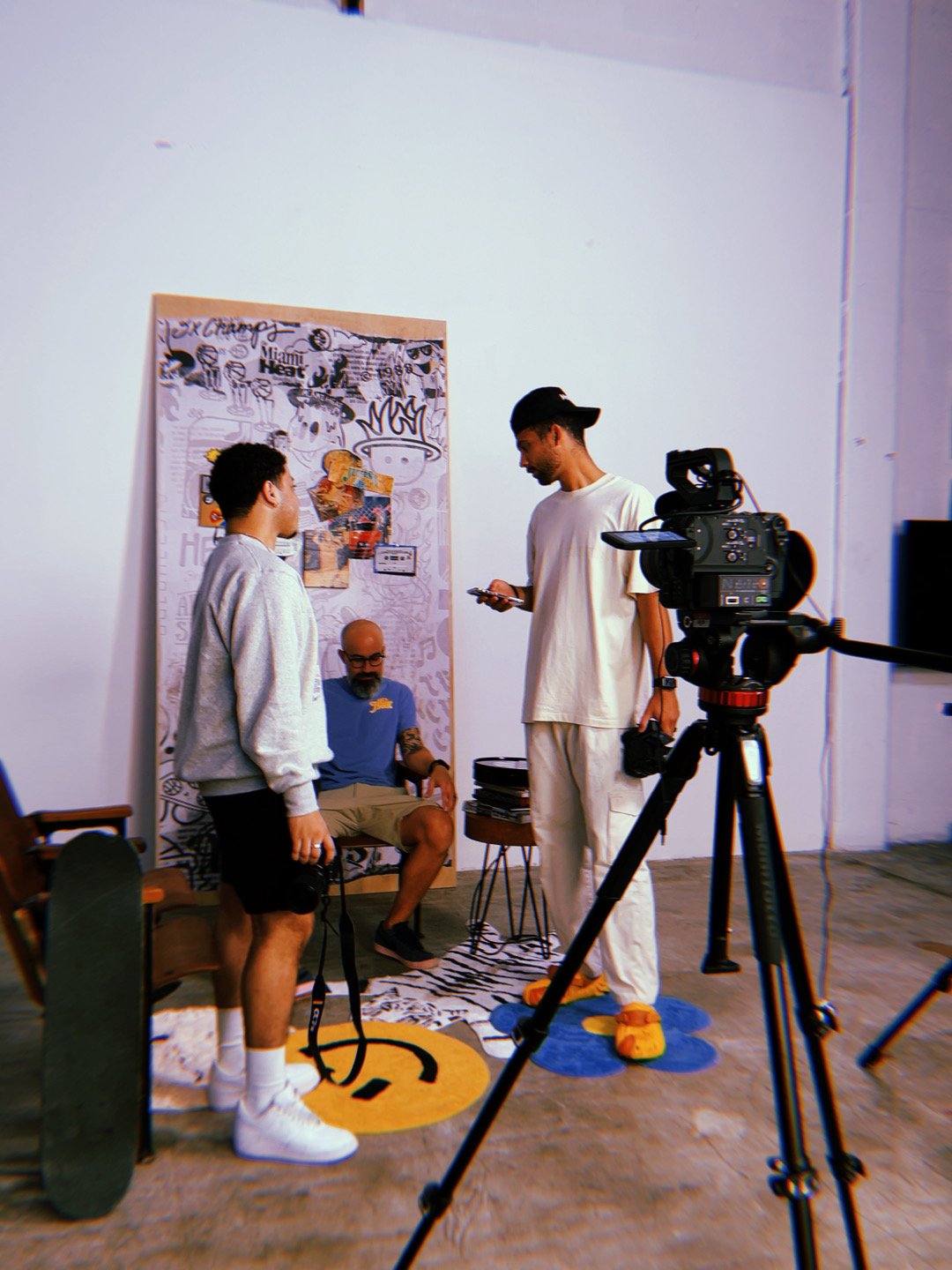 Three young men in a photography or video shoot setting, with one sitting in a chair and two standing, surrounded by camera equipment and artwork on a wall.