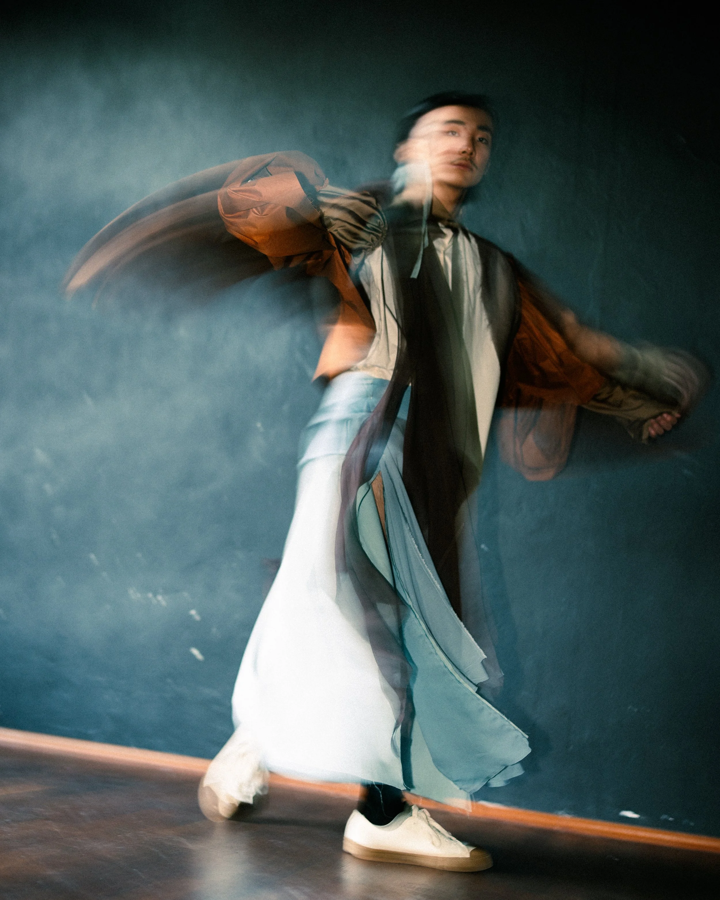 Motion-blurred photo of a young man in casual clothes and a jacket, standing against a dark wall.