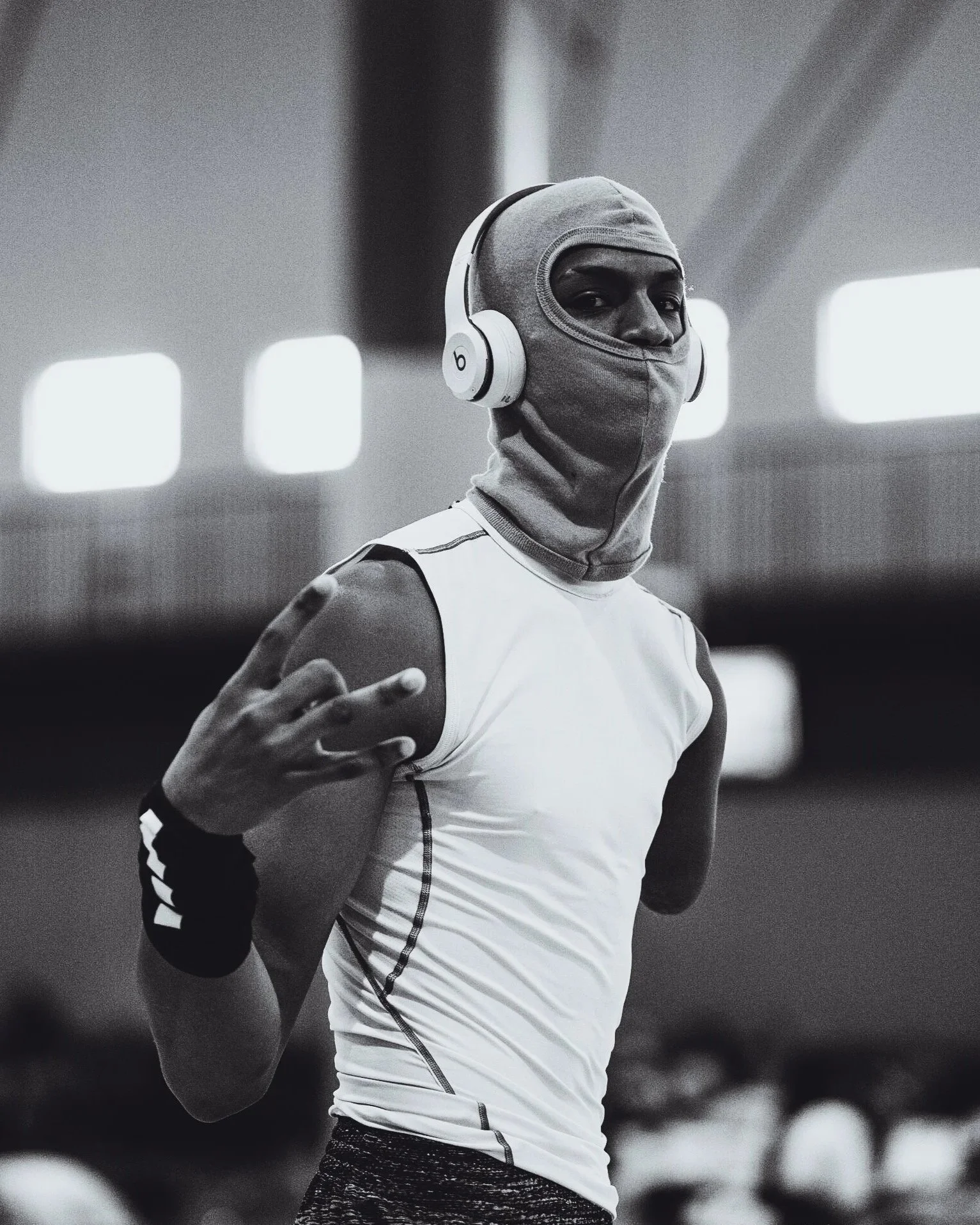 A person wearing headphones and a balaclava, making a peace sign gesture with their hand, in a gym or indoor sports facility.