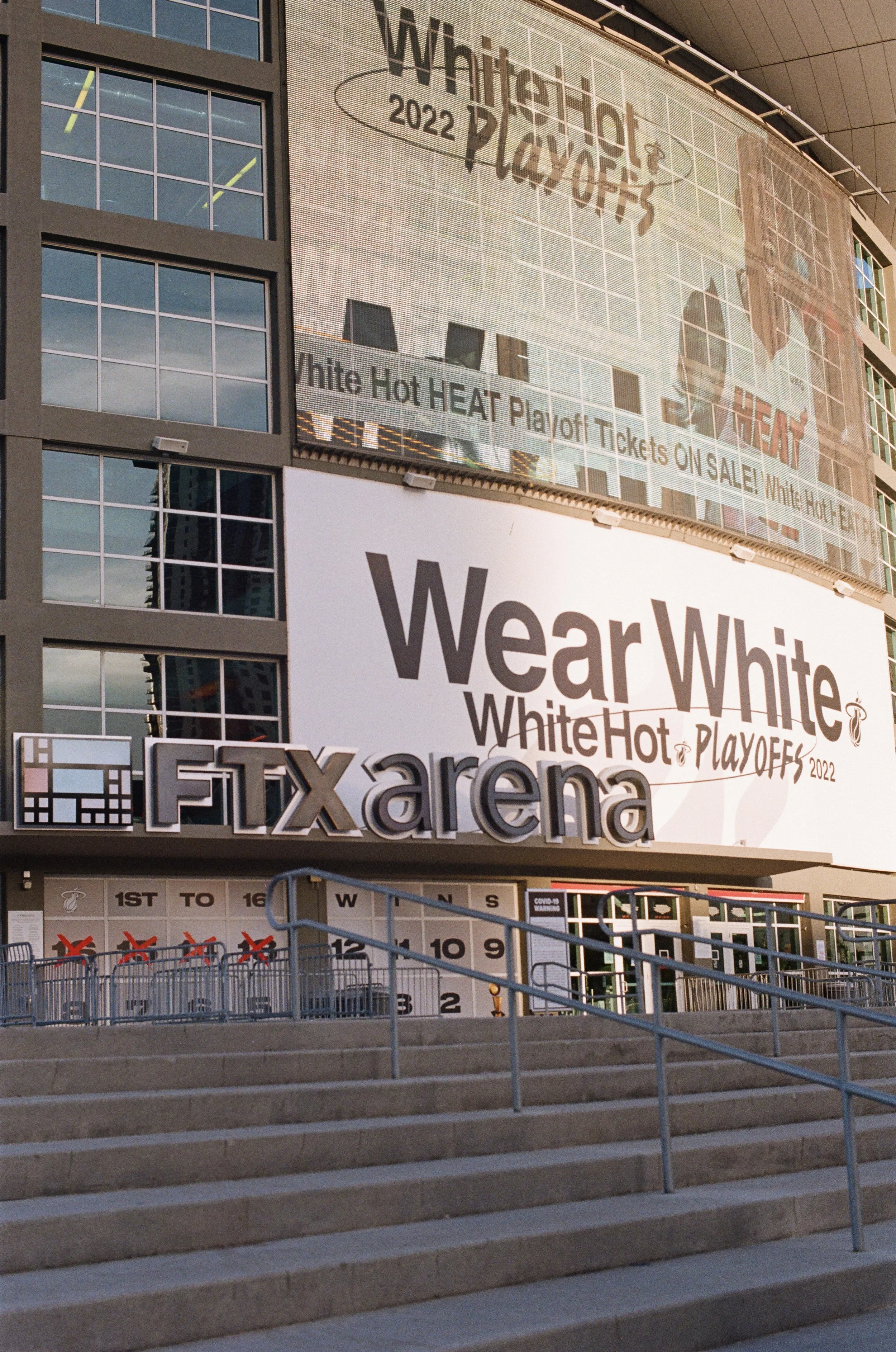 The exterior of the FTX Arena displaying large posters for the 2022 White Hot Playoffs, including signs about ticket sales and game schedule, next to a stepladder and metal railings at the entrance.