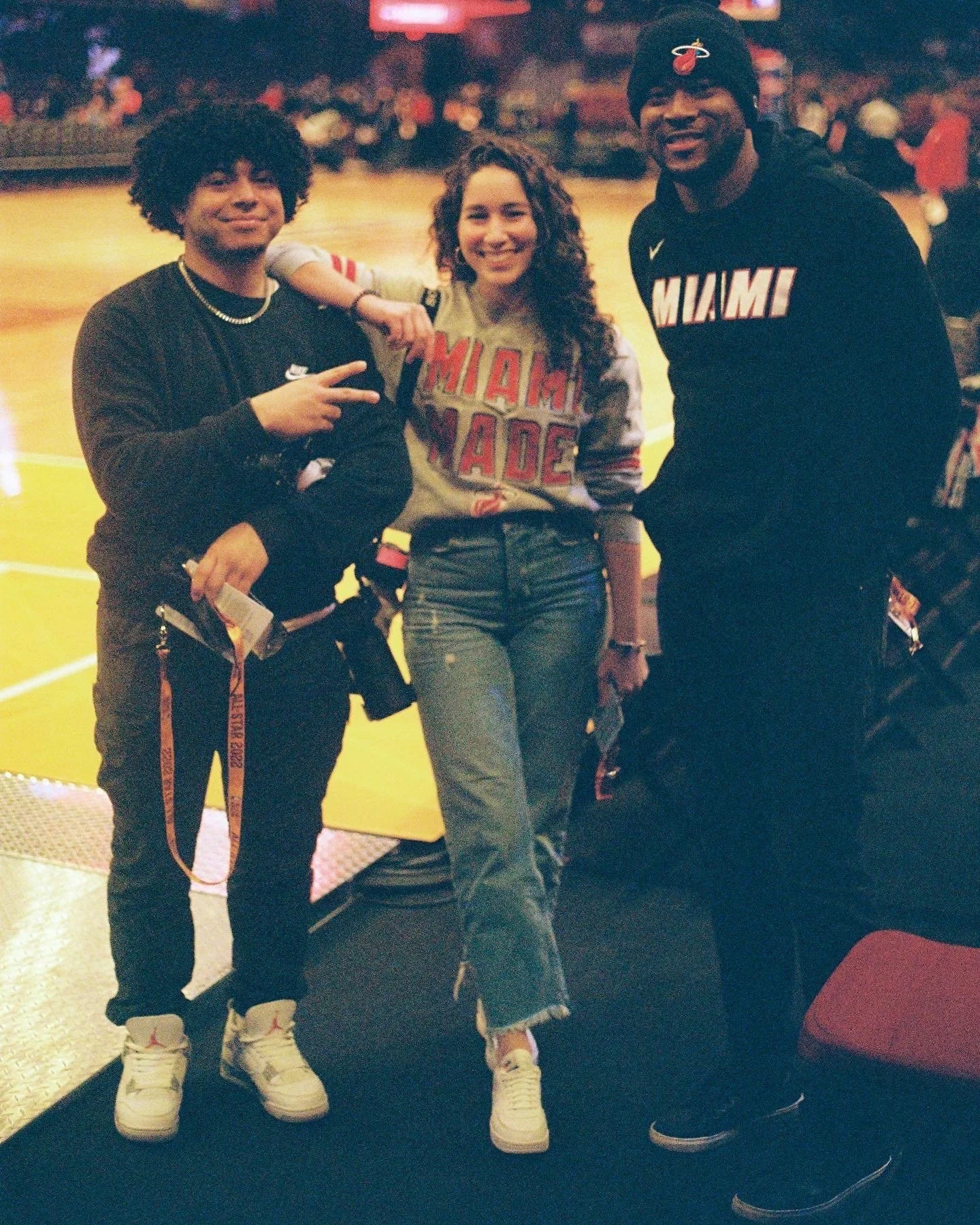 Three young people standing together in a basketball arena, smiling at the camera. The background features a basketball court and audience seating.