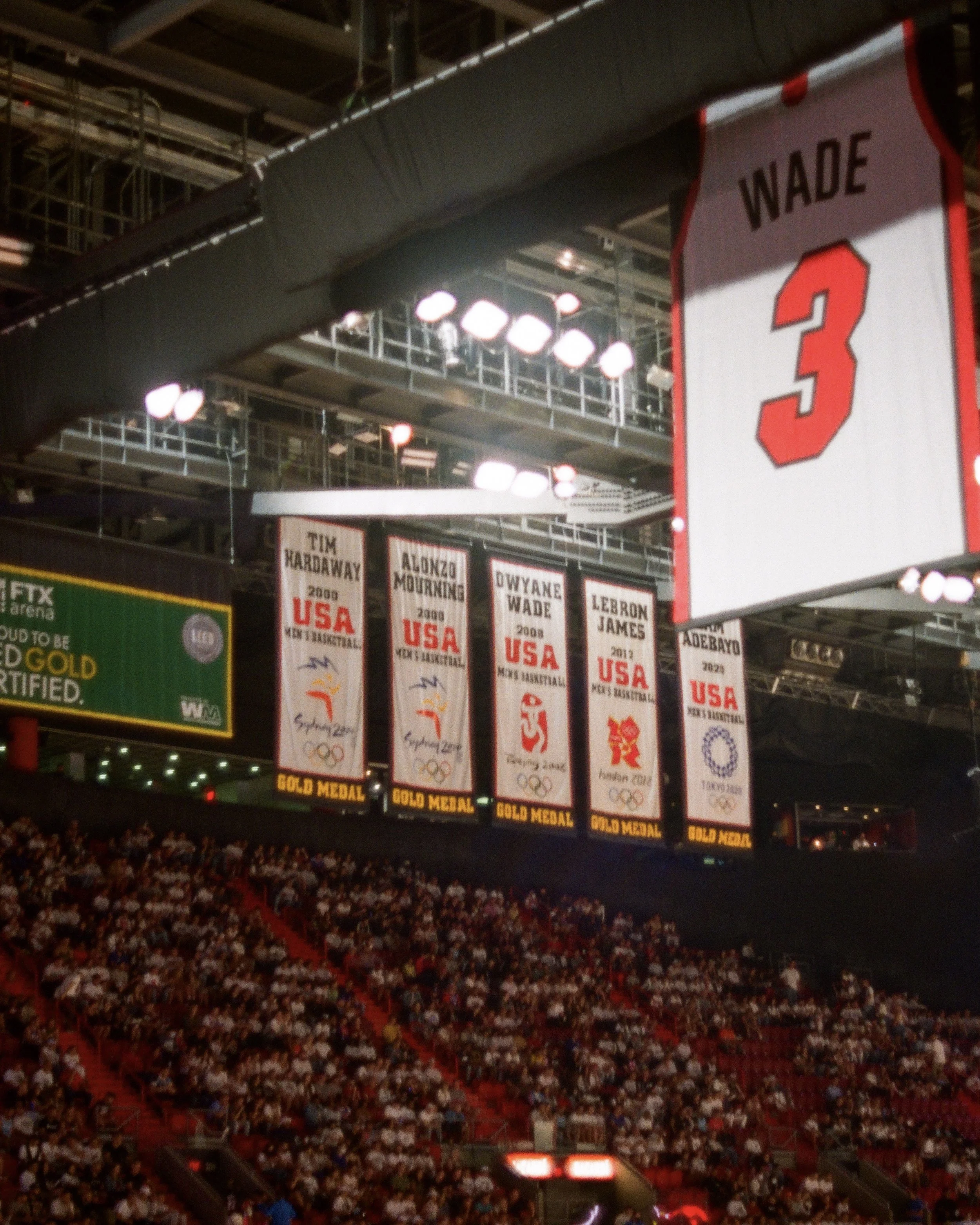 A basketball arena with banners hanging from the ceiling, including one for Wade with the number 3. The banners commemorate Olympic gold medals won by the USA basketball team, featuring players Tim Hardaway, Alonzo Mourning, Dwyane Wade, LeBron James