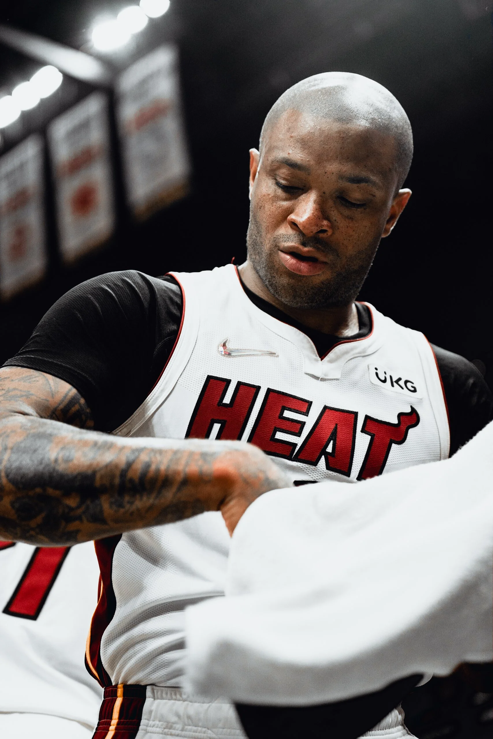 A basketball player with tattoos on his arms wearing a Miami Heat jersey, looking down and adjusting his clothing.