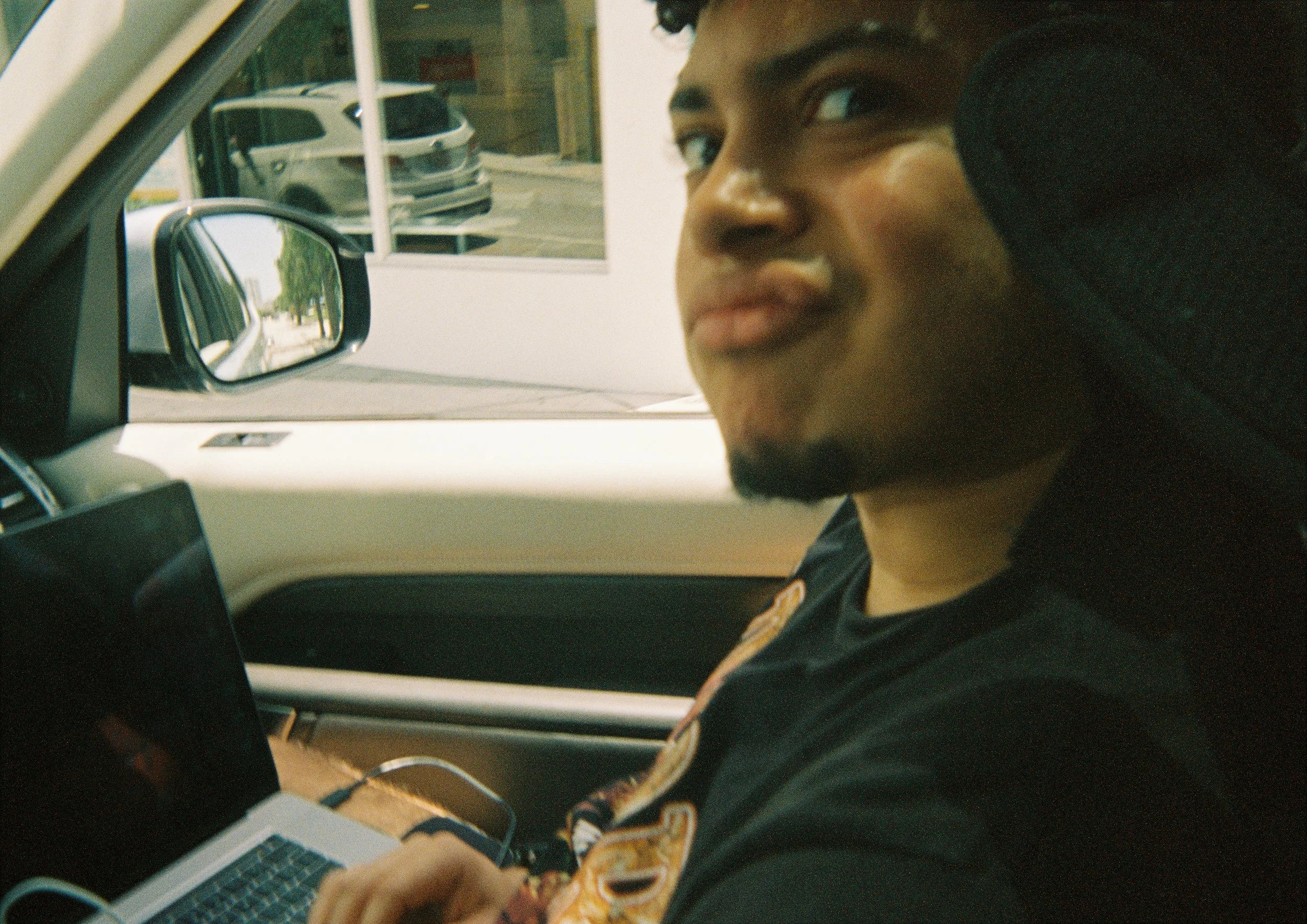 A young man sitting in a car, using a laptop on his lap. He is looking at the camera with a neutral expression. The car is parked, and the side mirror shows parked cars and a building outside.