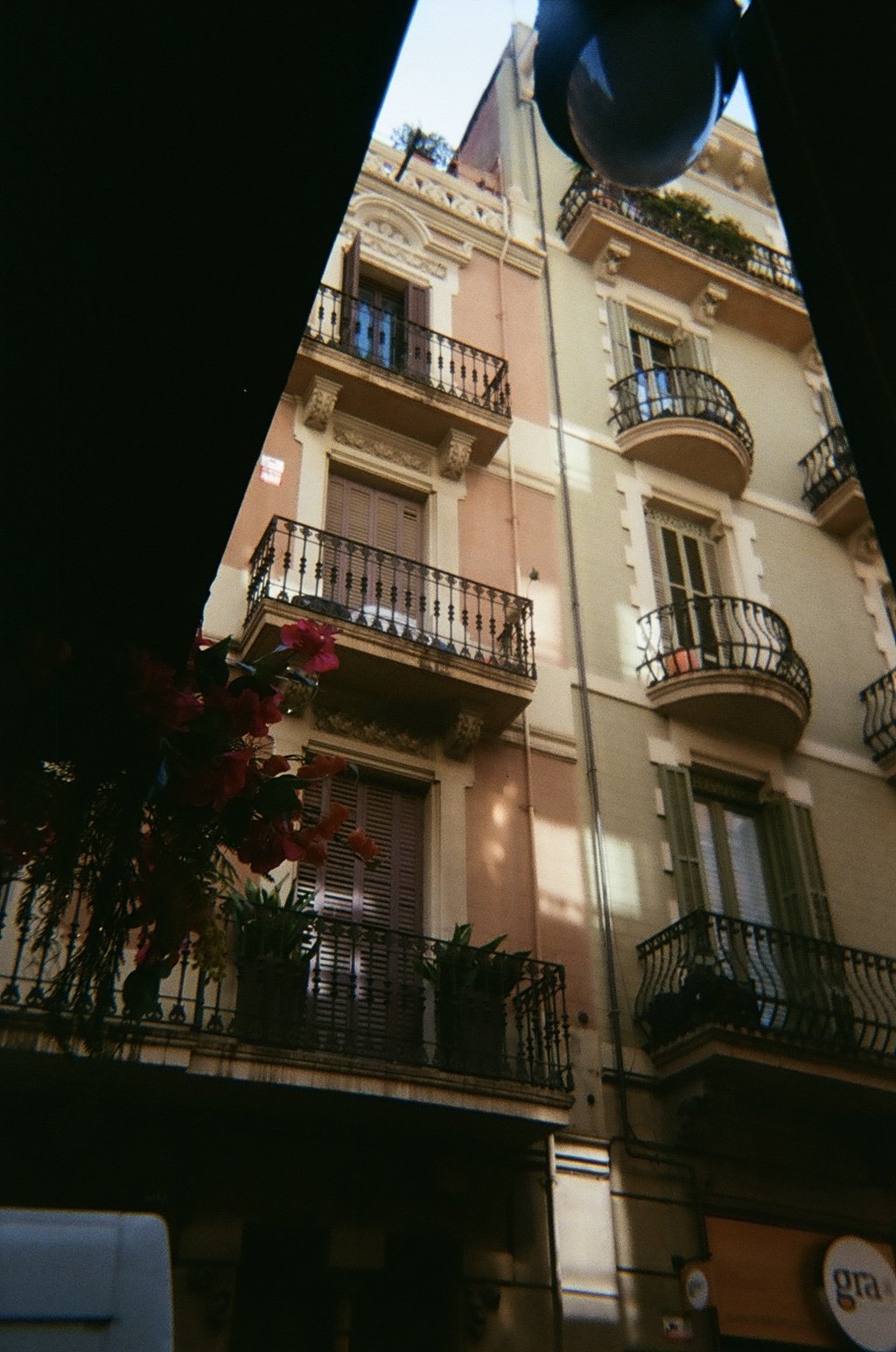 View of a multi-story building with curved balconies, some with potted plants, seen from below through a gap between dark structures, with a part of a street sign in the bottom right corner.